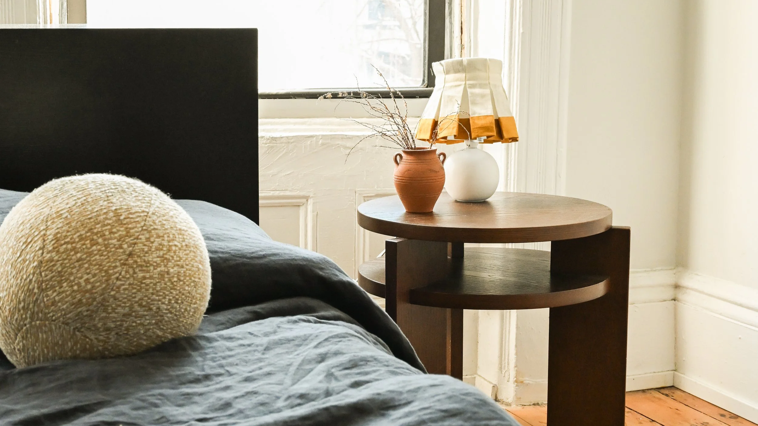 Close-up of a beige textured round pillow on a bed with dark gray bedding, bedside table with two vases, one terracotta and one white, with dried branches, near a window with sunlight streaming in.