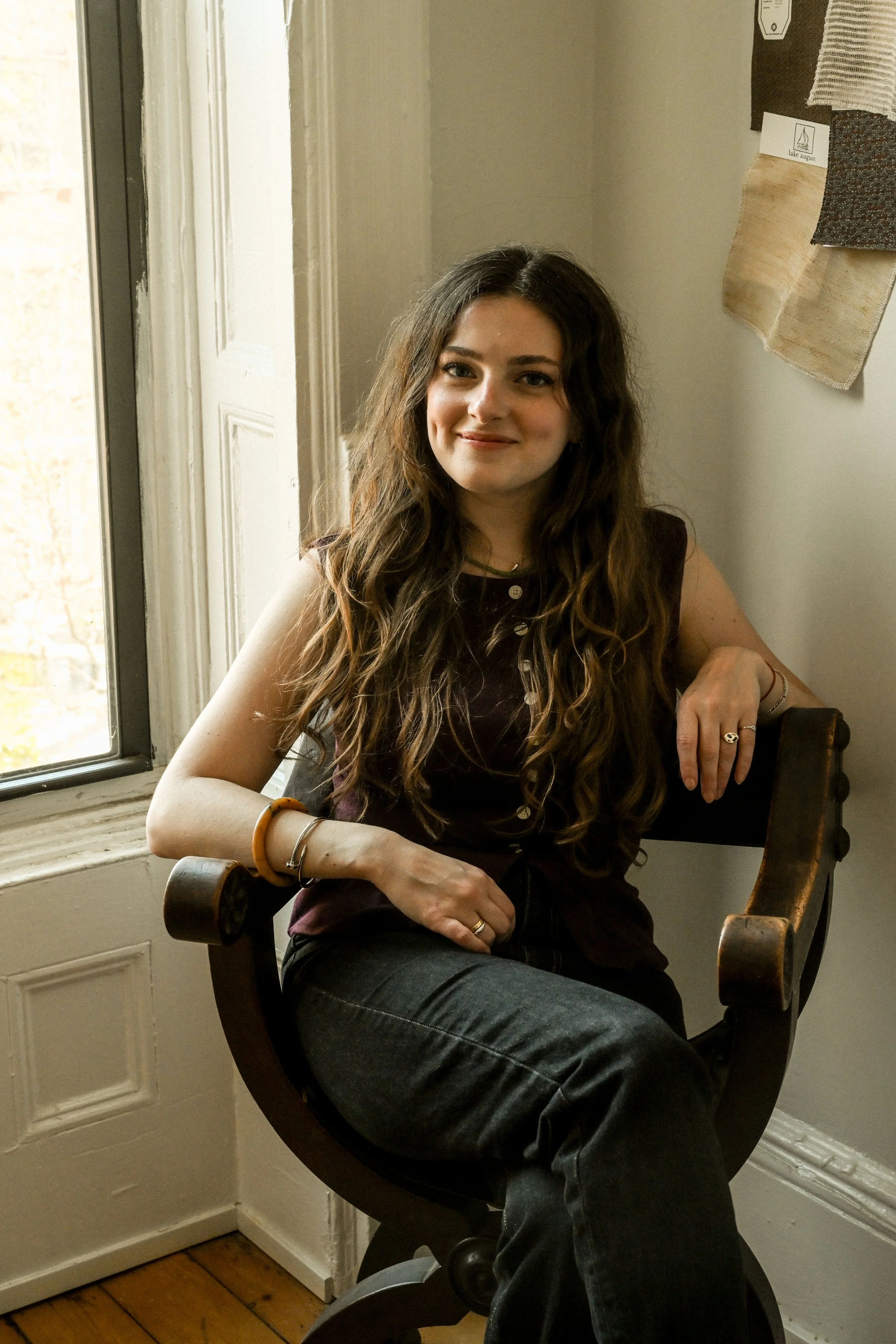 A young woman with long curly hair sitting on a rustic wooden chair by a window with light-colored walls and artwork on the wall.