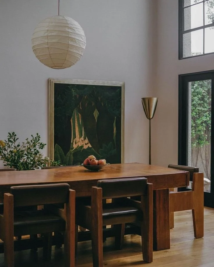 Interior of a dining room featuring a wooden table and chairs, a framed forest-themed artwork on the wall, a white paper lantern ceiling light, a gold floor lamp, and large window with a view of greenery.
