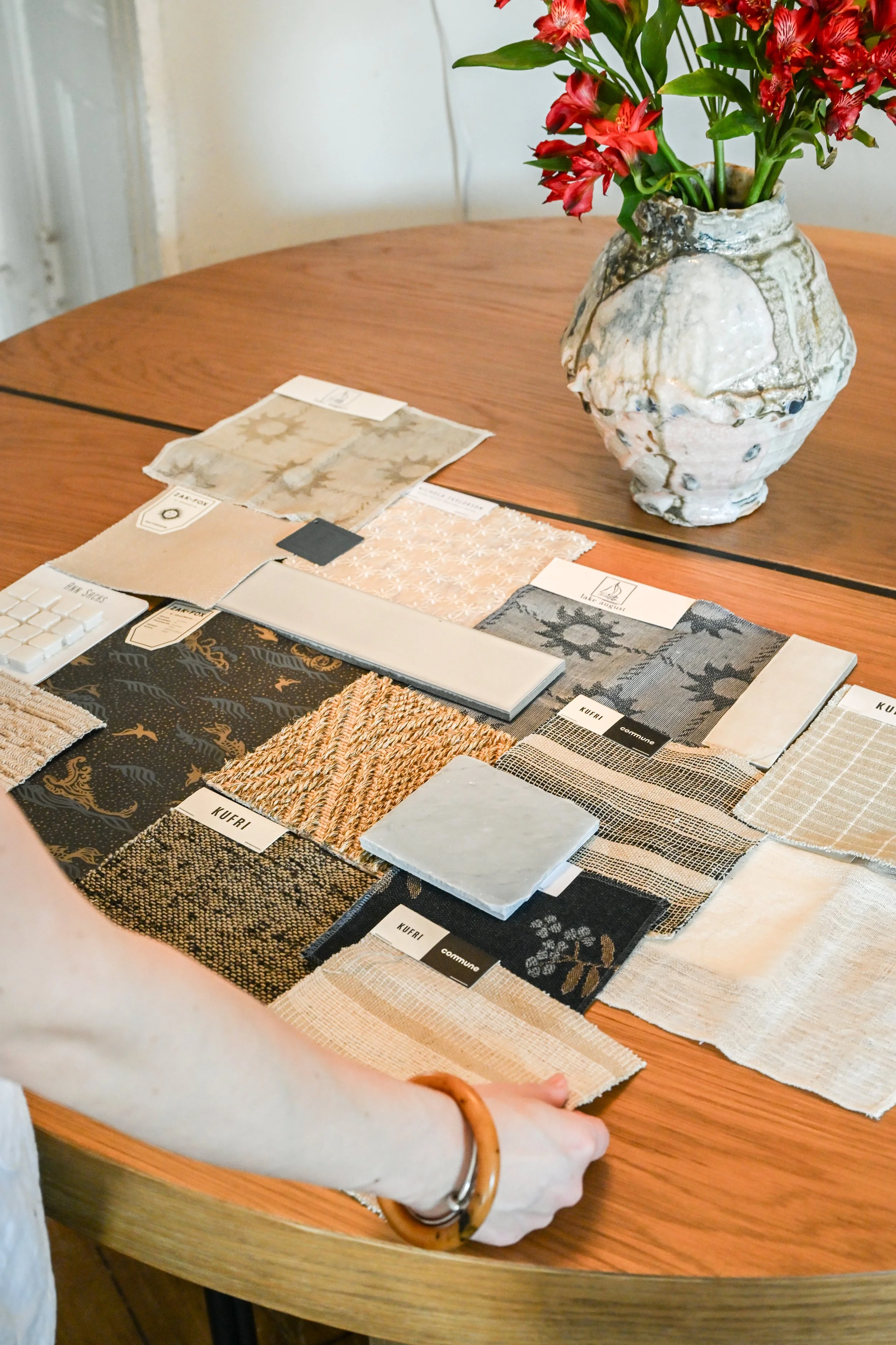 Person examining fabric swatches on a wooden table with a, textured ceramic vase filled with red flowers in the background.