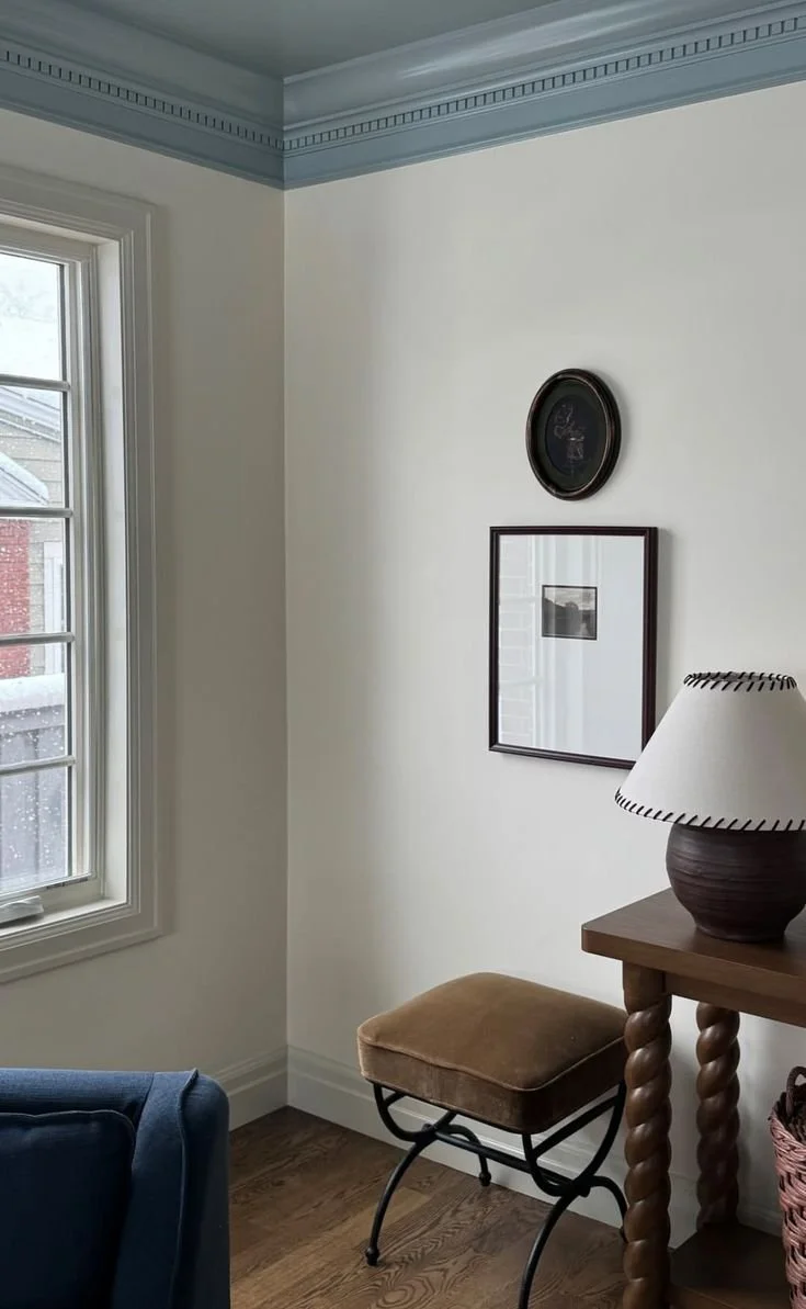 A corner of a living room with a window, wall art, a table lamp, and an upholstered footrest.