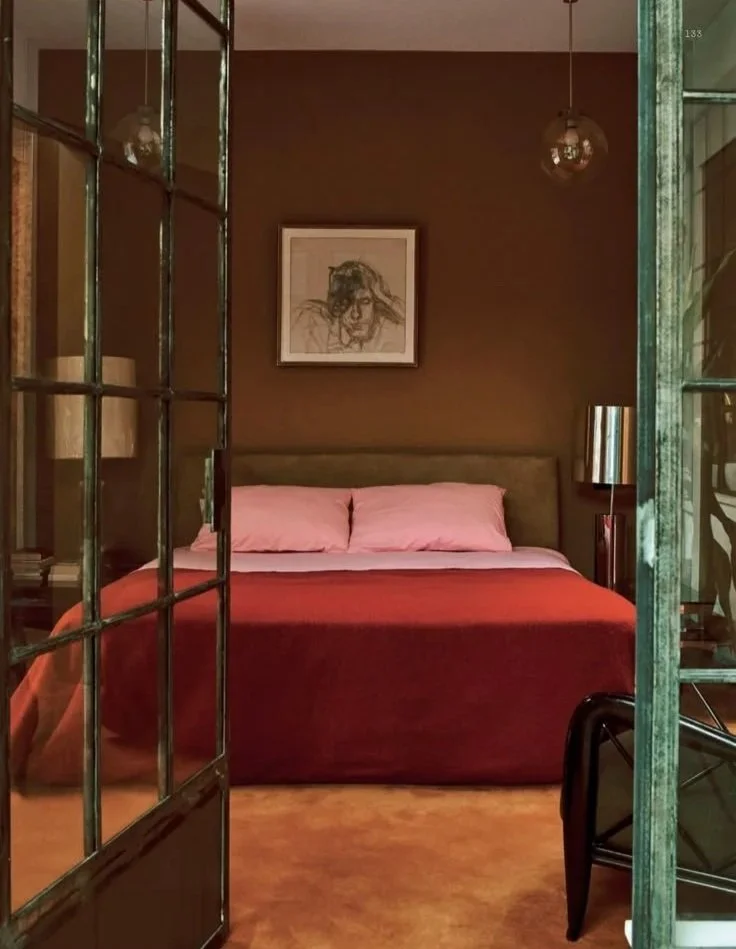 A bedroom seen through an open glass-paneled door with a dark brown wall, a framed sketch of a person's face above the bed, and pink pillows on the bed. There are glass lamps on each side of the bed and a small table with a lamp on the right side.