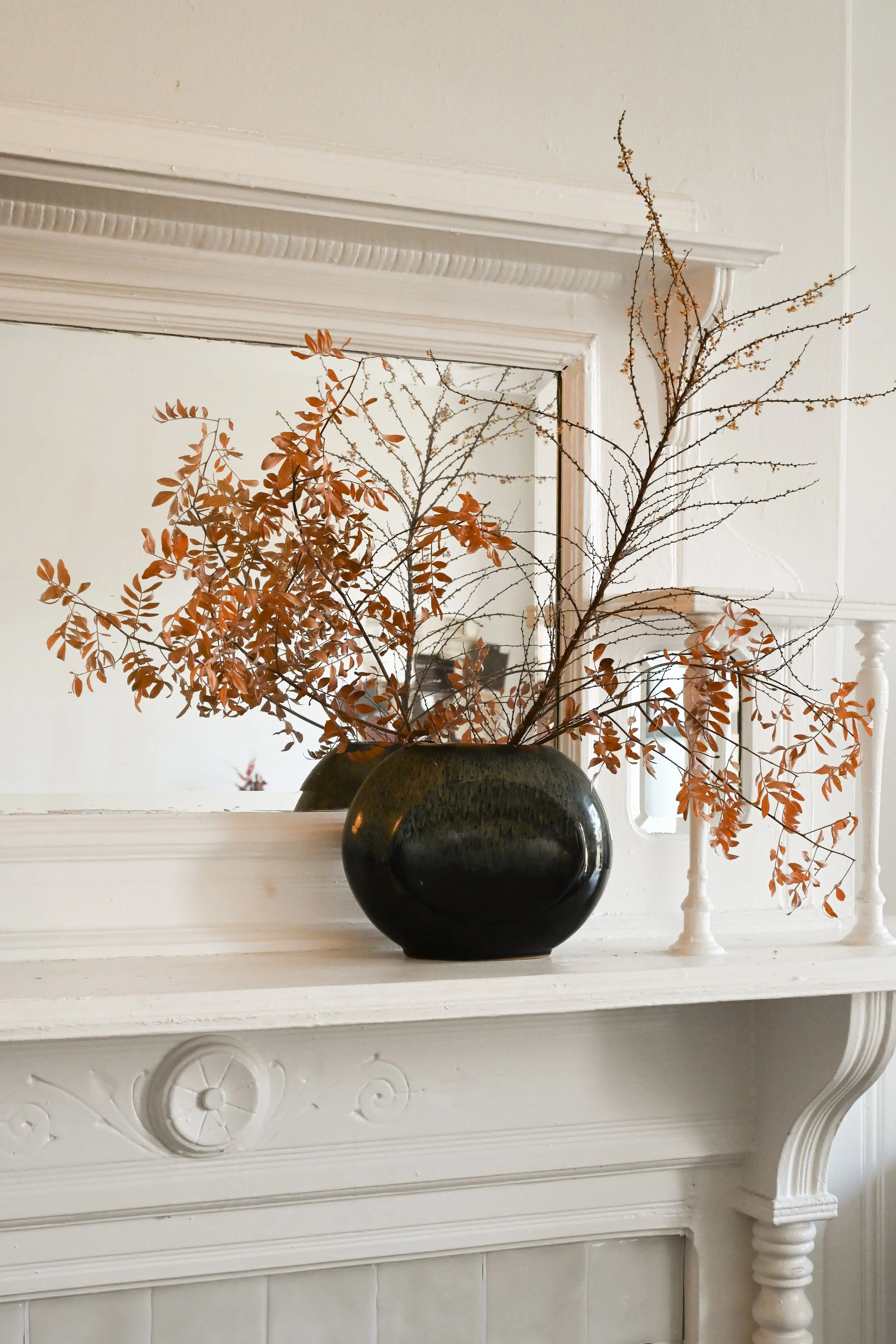 A decorative white fireplace mantel with intricate scrollwork, holding two vases filled with autumnal branches with brown leaves, against a white wall with a mirror reflecting additional branches.
