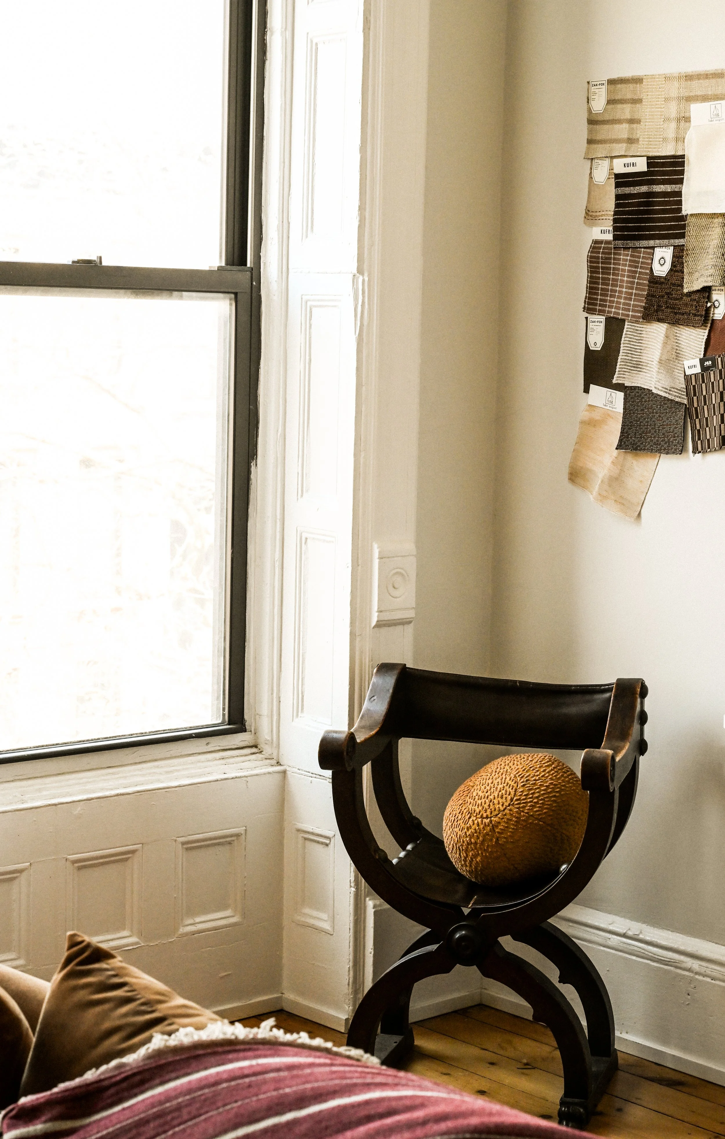 A corner of a room with a large window and wooden paneling. A black wooden chair with a round, textured cushion on it is near the window. On the wall, fabric swatches are pinned, showing various colors and patterns.