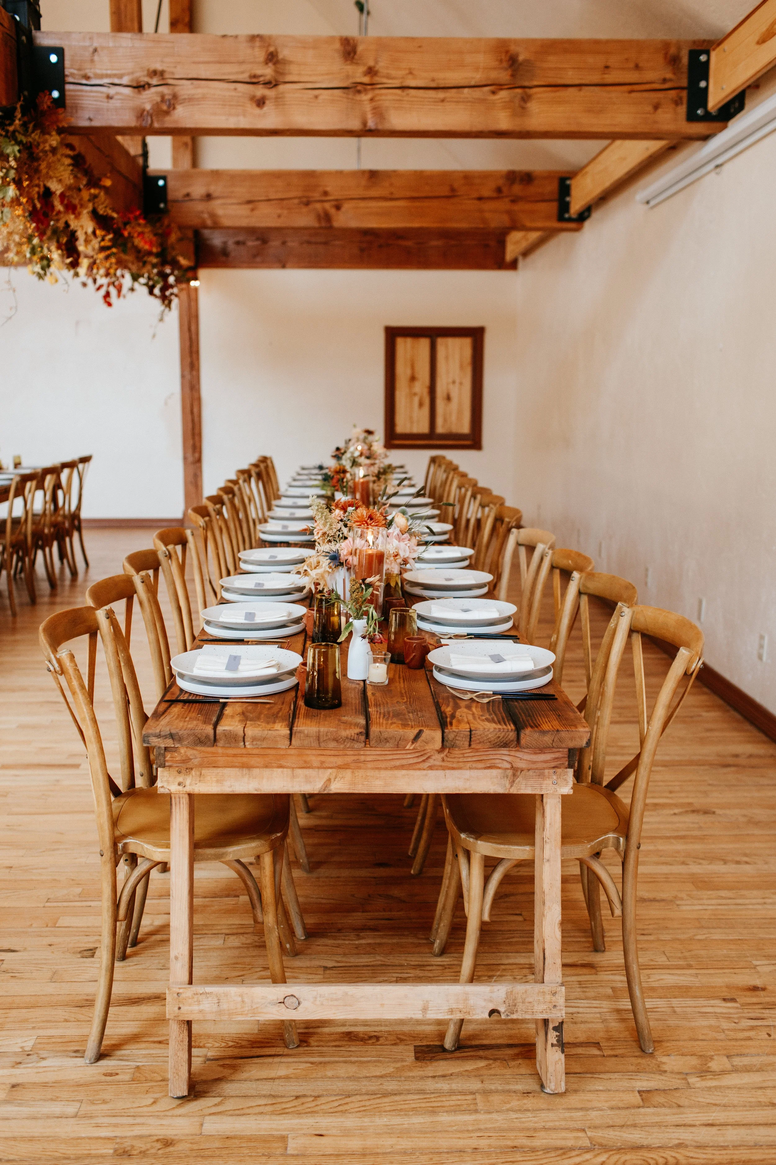 Long wooden dining table set with white plates, gold and amber glasses, and pink and orange floral centerpieces, in a rustic indoor space with wooden beams and a light-colored wall.