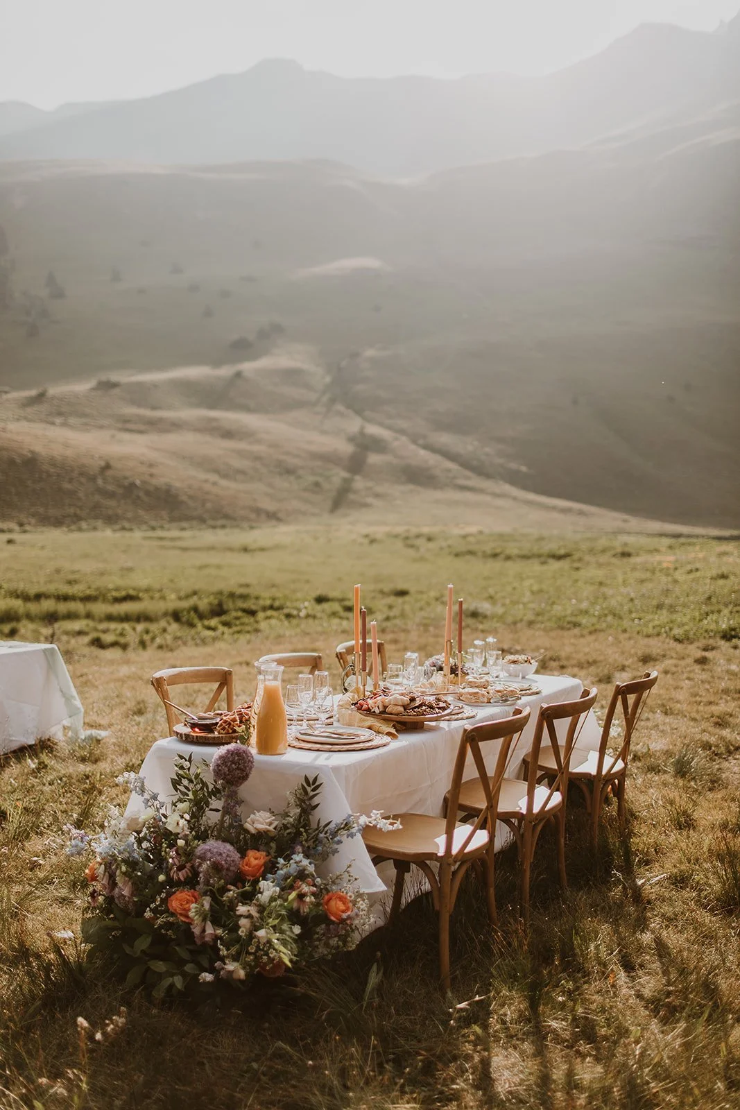 A set outdoor dining table with a white tablecloth, floral centerpiece, pink candles, and place settings, situated in a grassy field with rolling hills in the background.