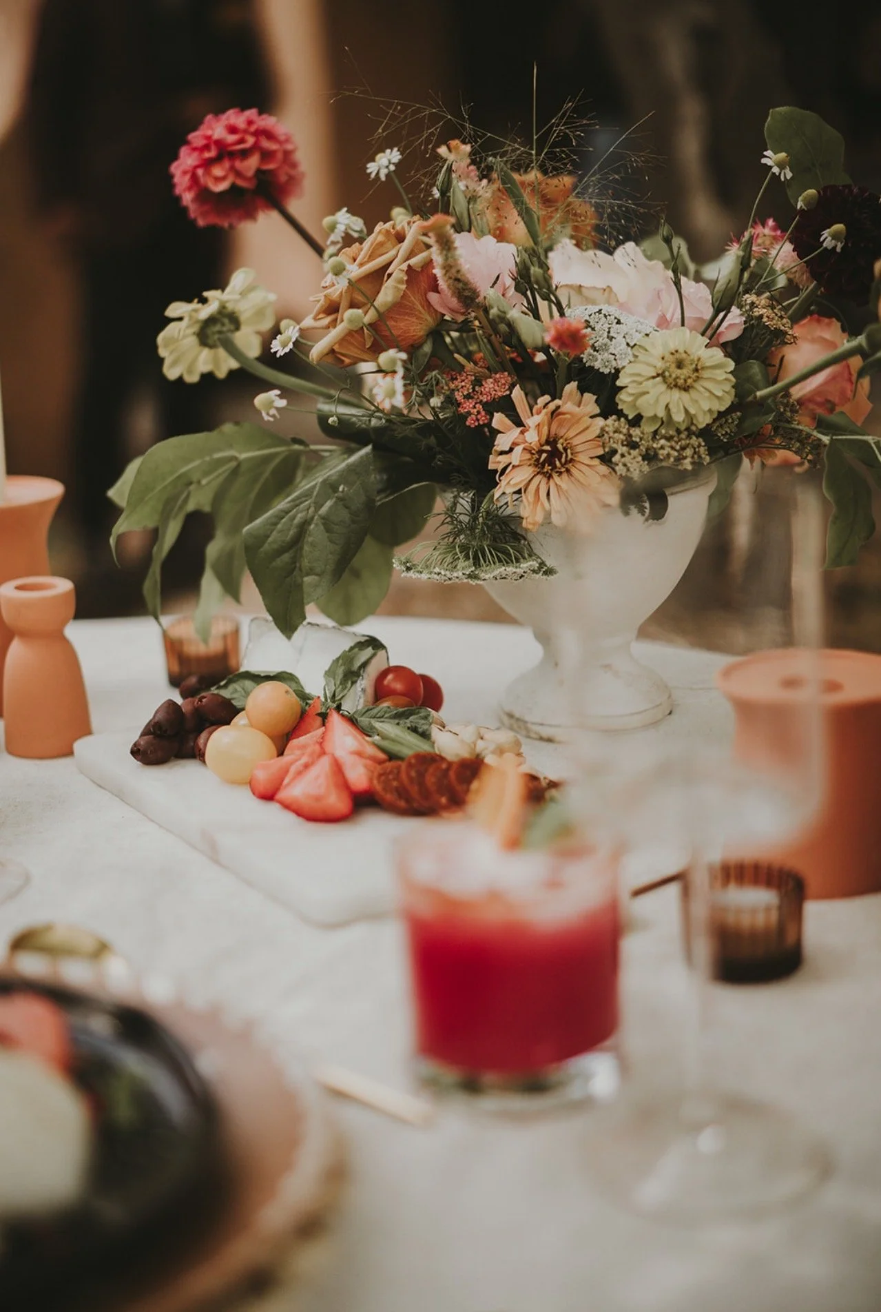 A floral centerpiece on a table with a variety of foods, including grapes, watermelon slices, cherry tomatoes, and a plate of charcuterie, along with candles and pottery against a blurred background.