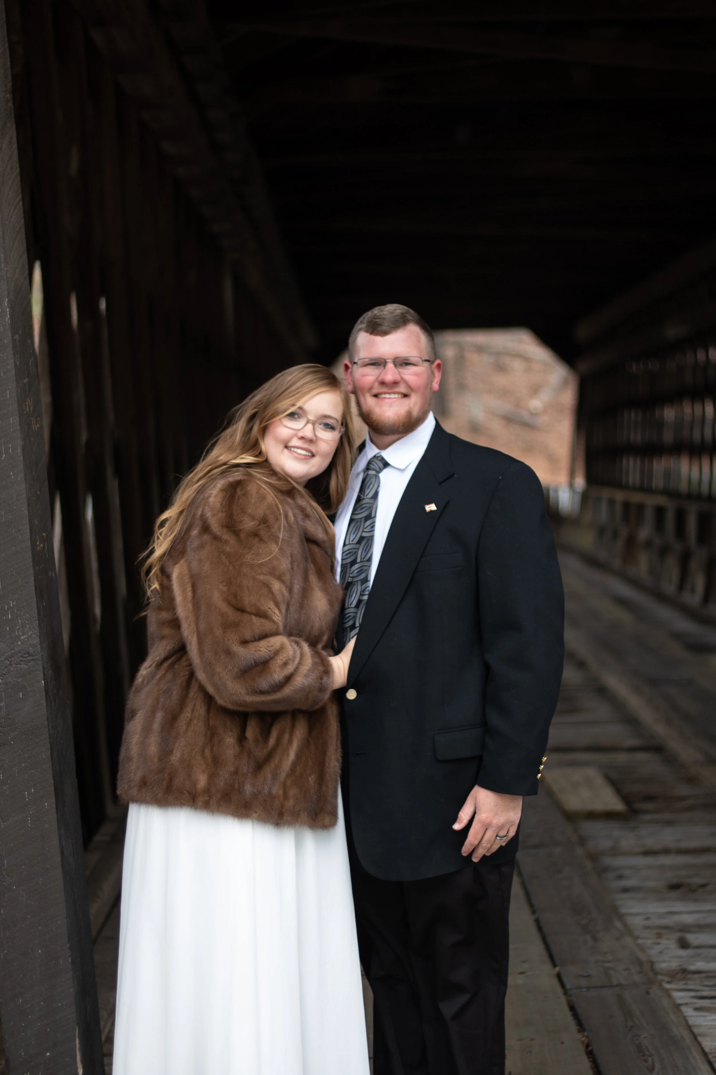 A couple dressed in formal attire standing on a wooden bridge, with the woman wearing a fur coat and white dress, and the man in a suit, smiling at the camera.