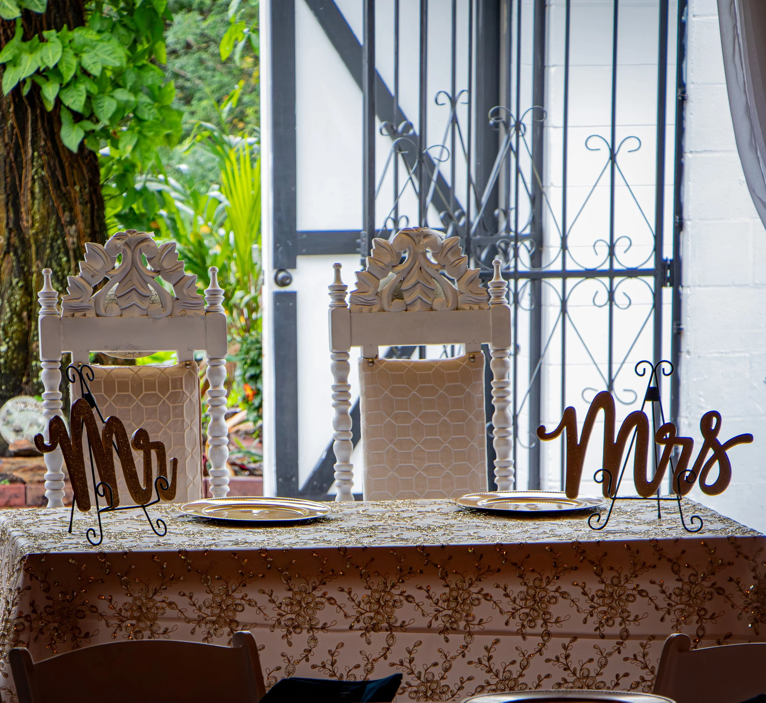 Decorated table set for a wedding or special celebration with 'Mrs' and 'Mr' signs, plates, and ornate chairs, outdoors in a garden or patio area.