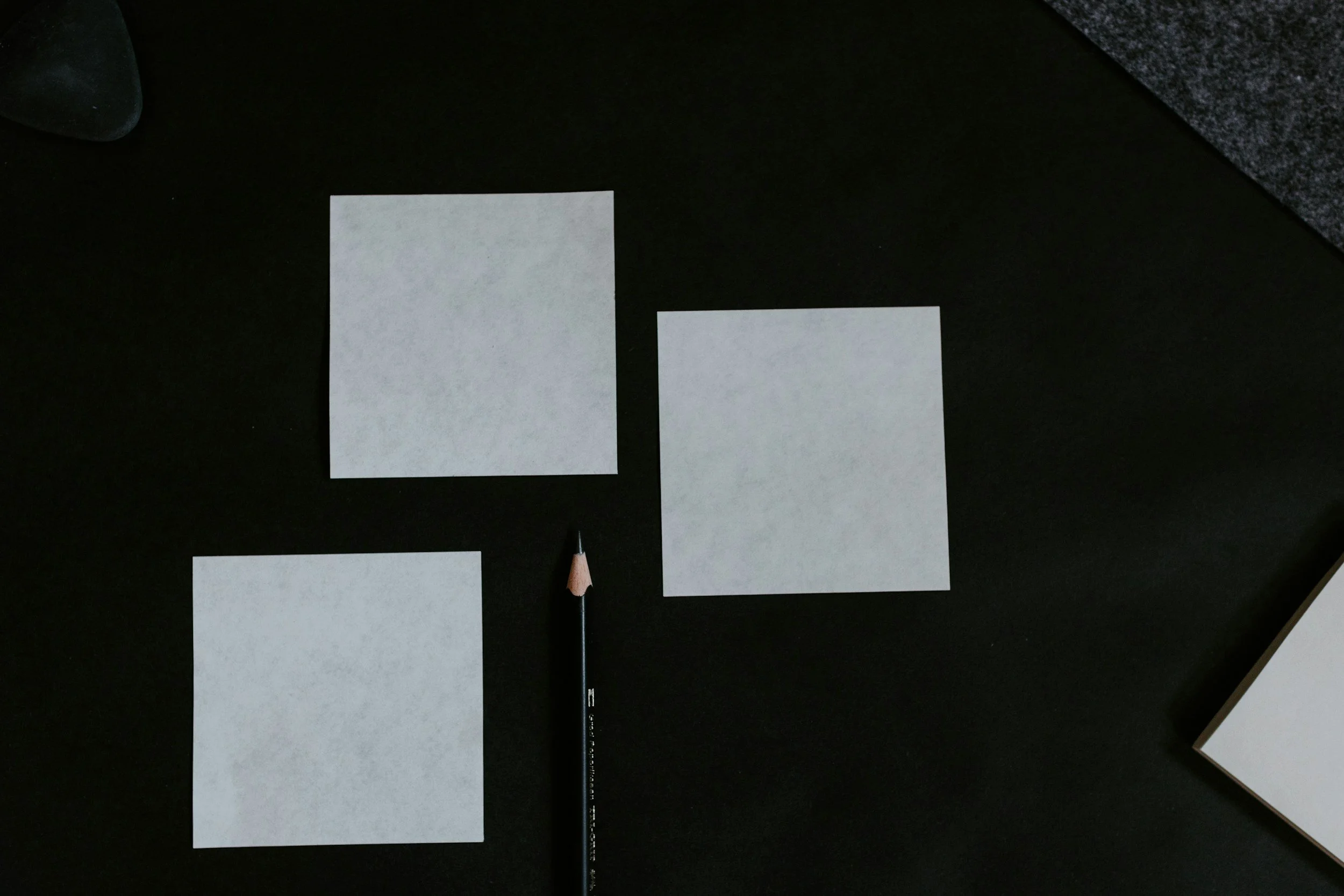 Three blank white square papers arranged on a black surface, with a black pencil positioned between them, and a dark textured item in the upper right corner.