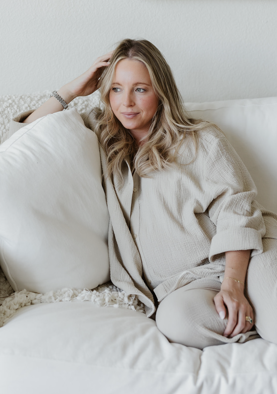 A woman with blonde hair sitting on a white sofa, resting her head on a pillow, wearing a beige shirt and light-colored pants.
