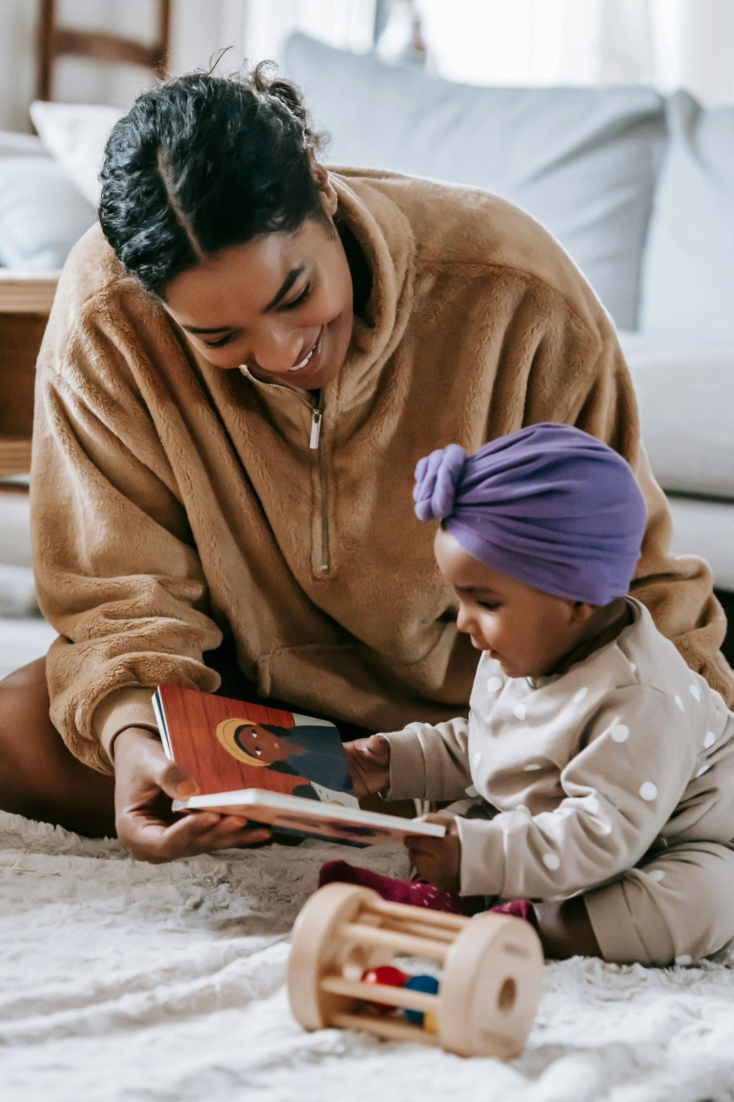A woman and a young girl sitting on a plush carpet in a cozy living room, looking at a children's book together with a wooden toy in the foreground.