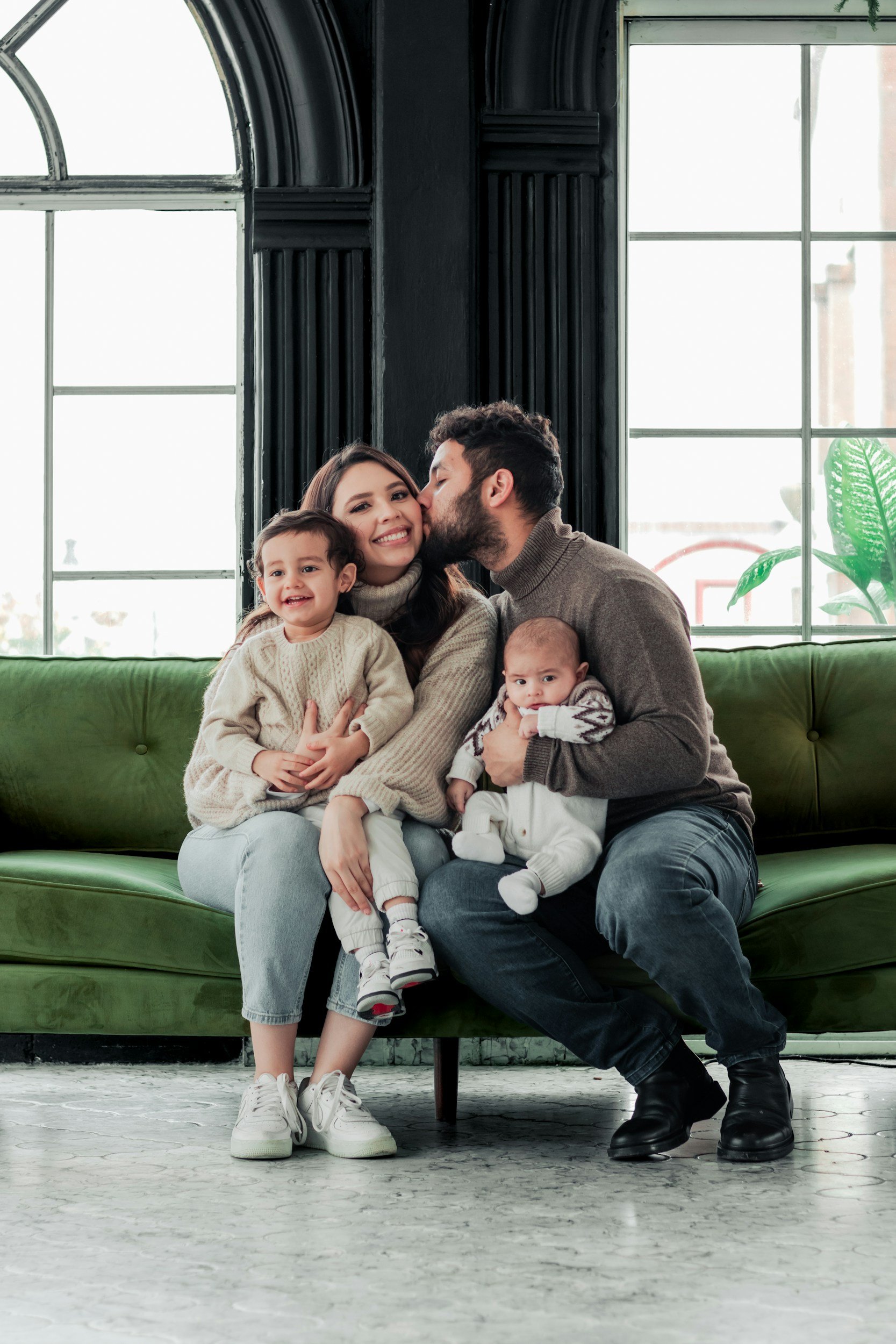 A family of four sitting on a green couch in front of large windows. The family includes a smiling woman, a man with a beard kissing her cheek, a young girl with curly hair sitting on the woman's lap, and a baby sitting on the man's lap. The room has black walls, and there is a plant with large green leaves near the window.