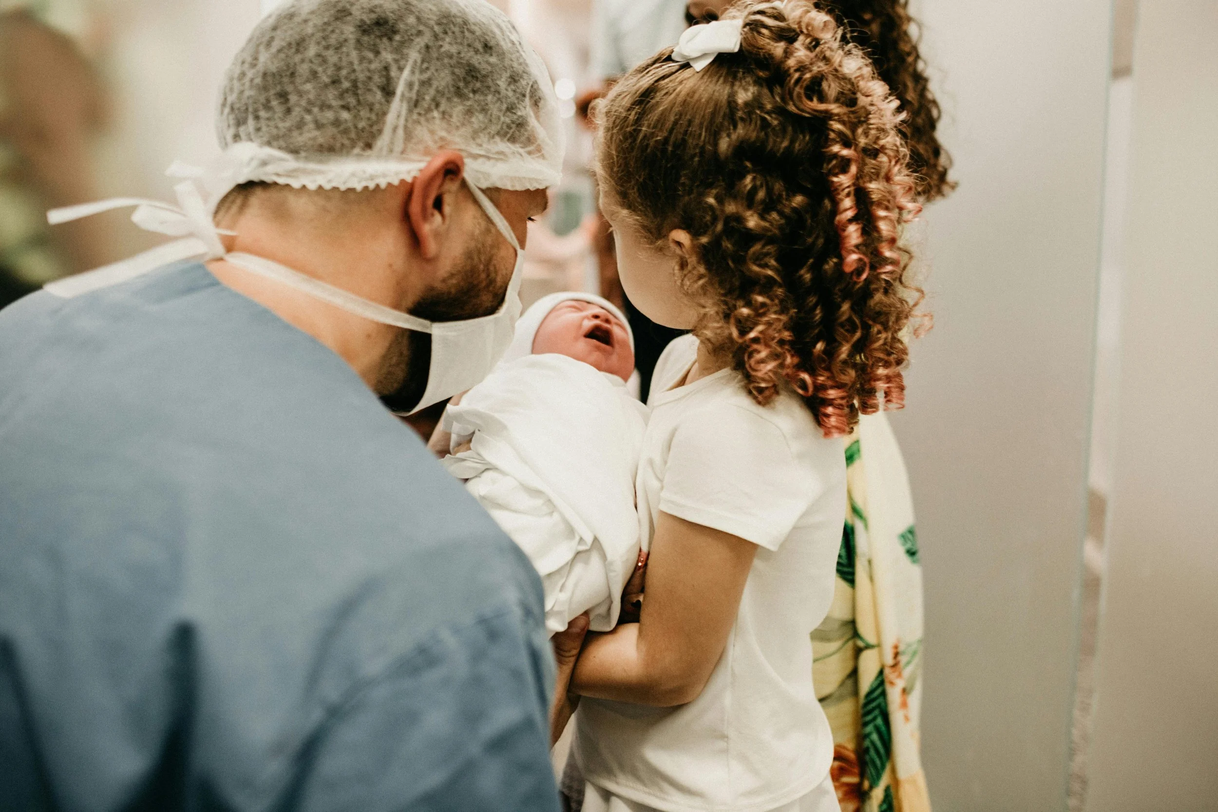 A hospital scene with a man, woman, and a newborn baby. The man is wearing scrubs, a surgical mask, and a hair cover, while the woman is holding the baby, who is dressed in white. The baby is crying, and the woman has curly hair with reddish highlights. The group is close together, sharing a special moment.