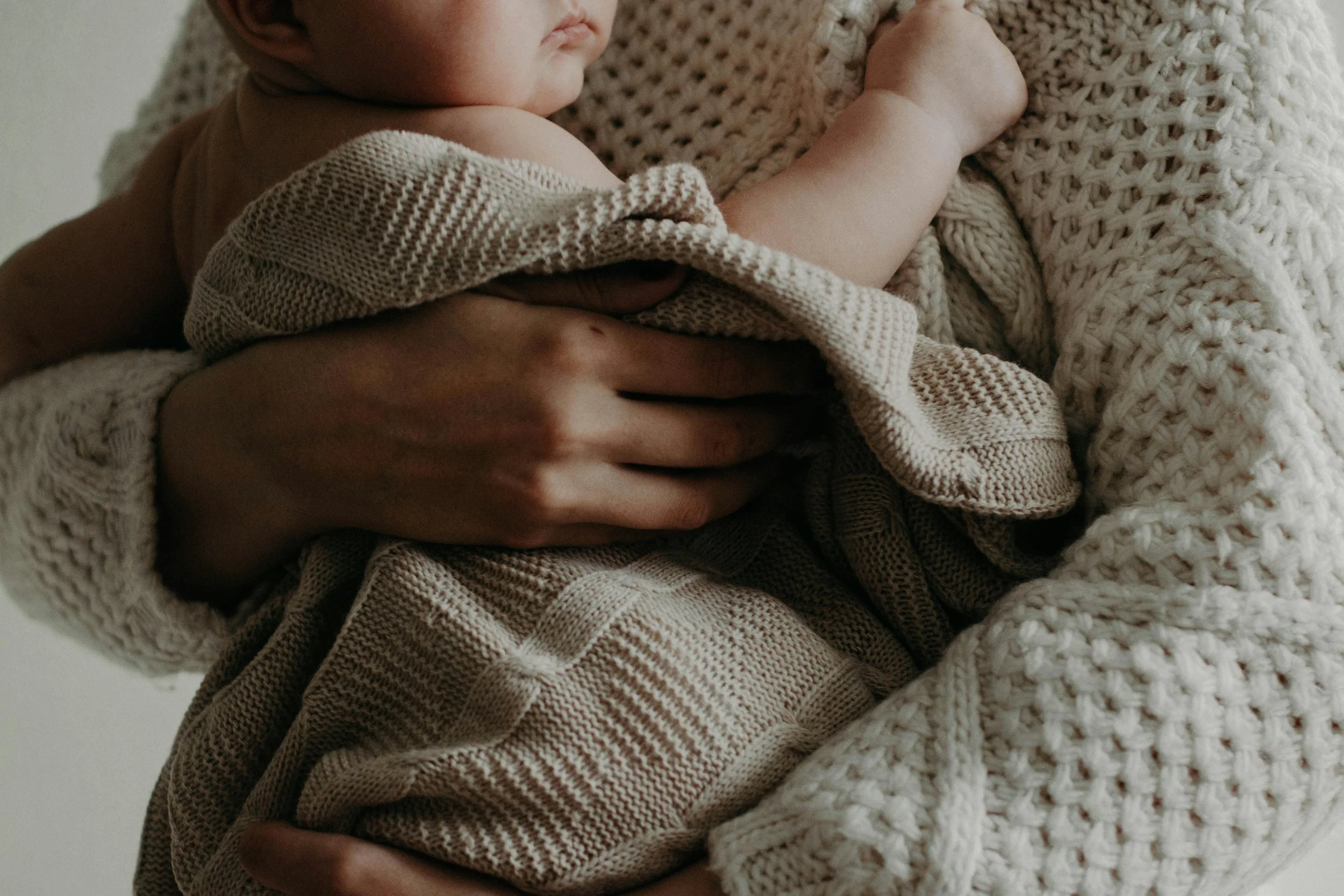 A close-up of a baby being held in someone's arms, wrapped in knitted beige and brown blankets, with part of the baby's face and body visible.