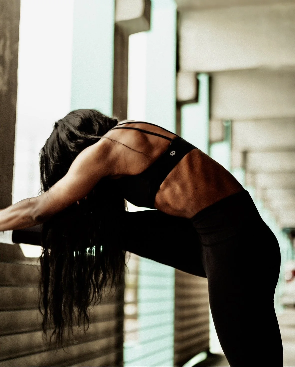 A woman with long dark hair doing a forward bend stretch, leaning over a wooden surface in a gym or fitness studio.