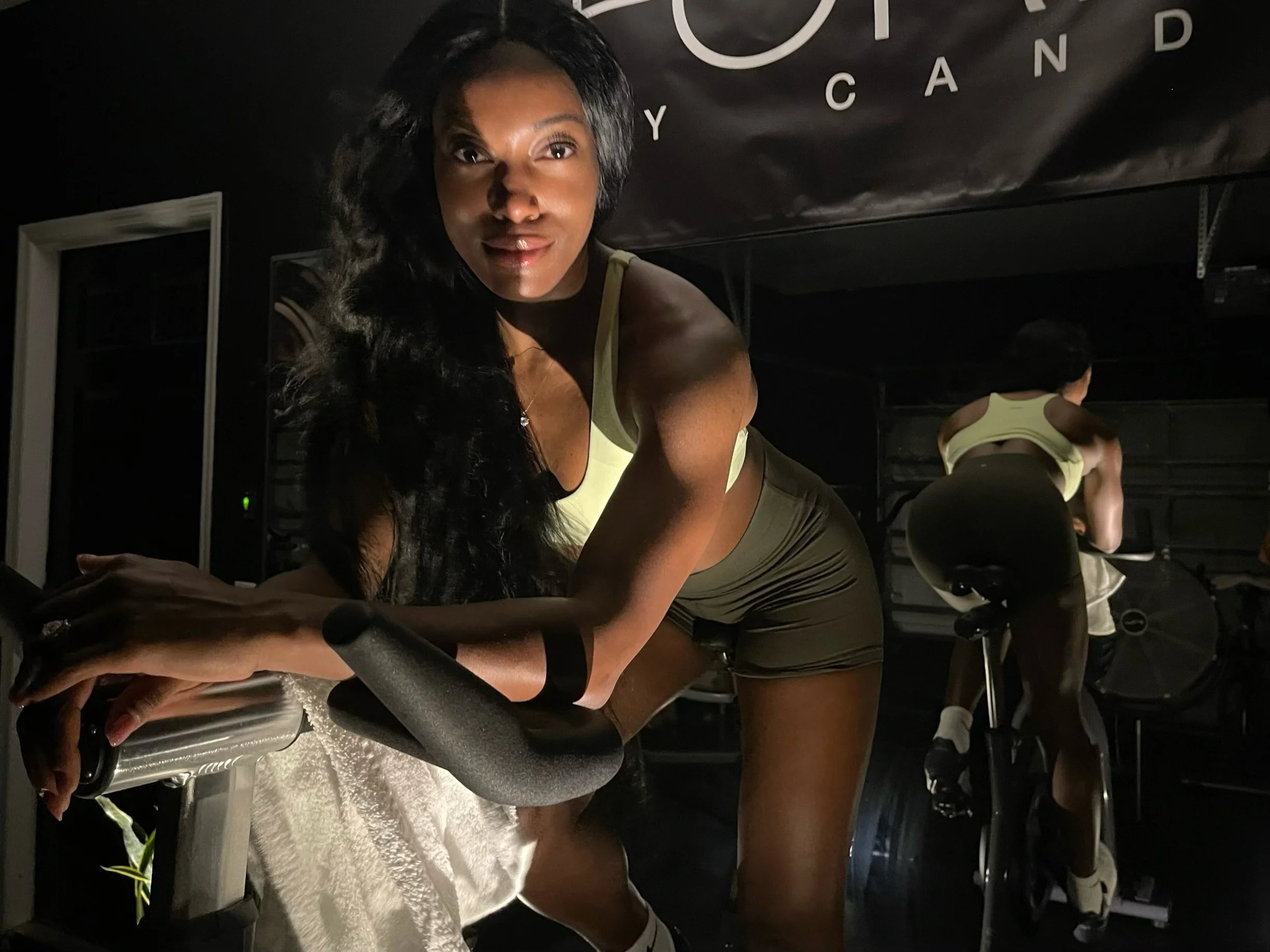 Woman with long black hair and athletic build leaning on exercise bike in a dimly lit gym, with another person on a stationary bike in the background.