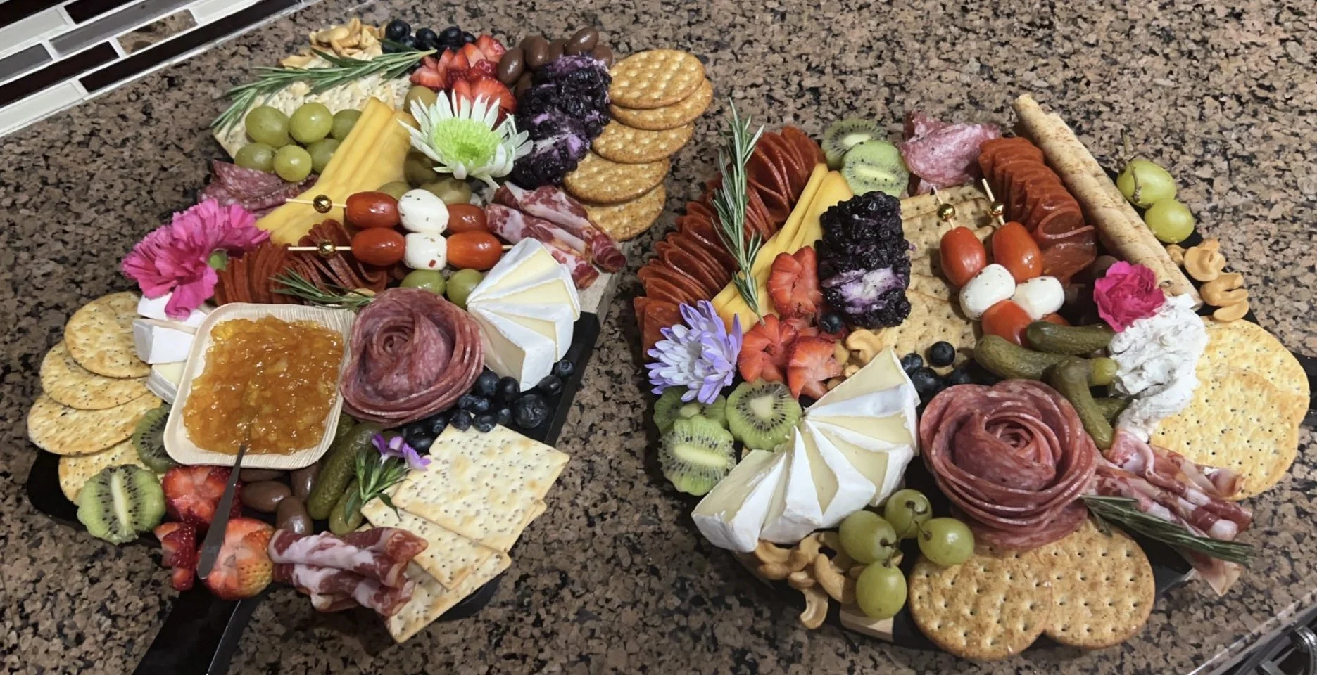 Charcuterie boards with assorted meats, cheeses, crackers, fruits, jams, and decorative flowers on a granite countertop.