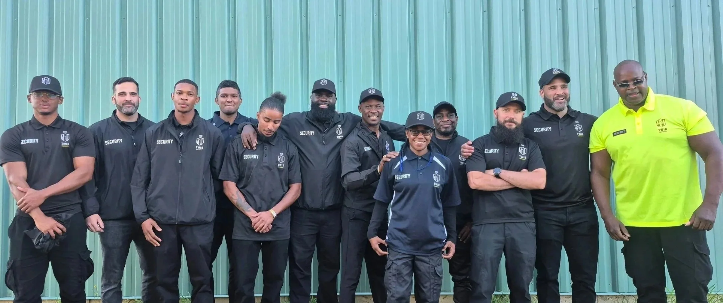 Group of security personnel standing in front of a green metal wall, wearing uniforms and caps with the REG Twin Security logo, one person on the far right in a lime green polo shirt among others.