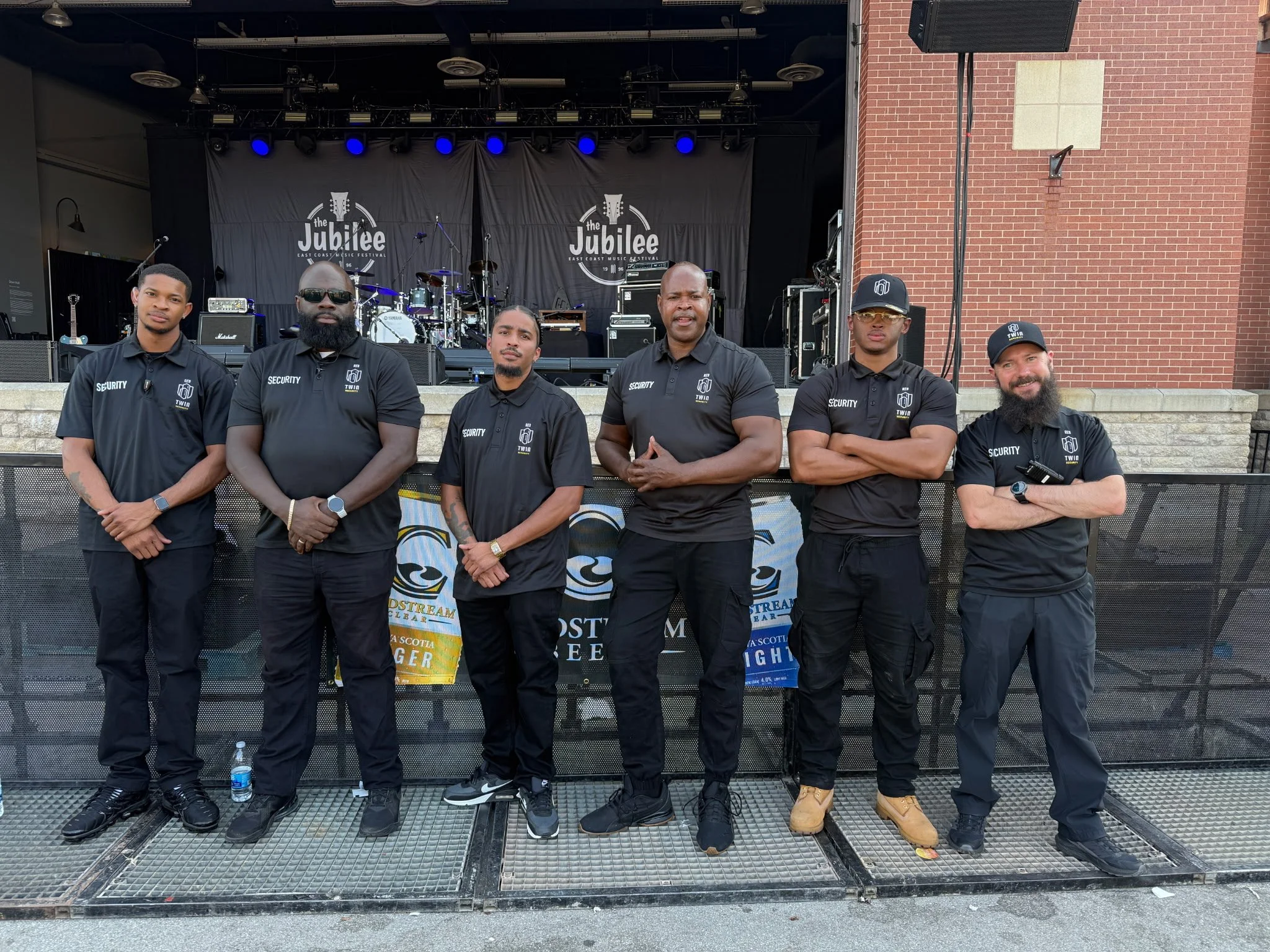 Six security personnel standing in front of a stage with musical instruments, wearing black shirts with security badges and posing for a photo before a concert.