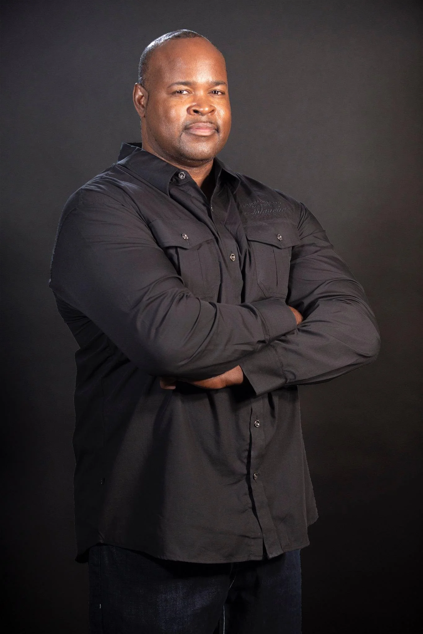 Richard Gray with crossed arms, wearing a black button-up shirt, standing against a dark background.