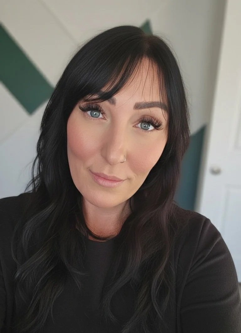 A woman with long black hair, blue eyes, and light makeup, wearing a black top, taking a selfie indoors.