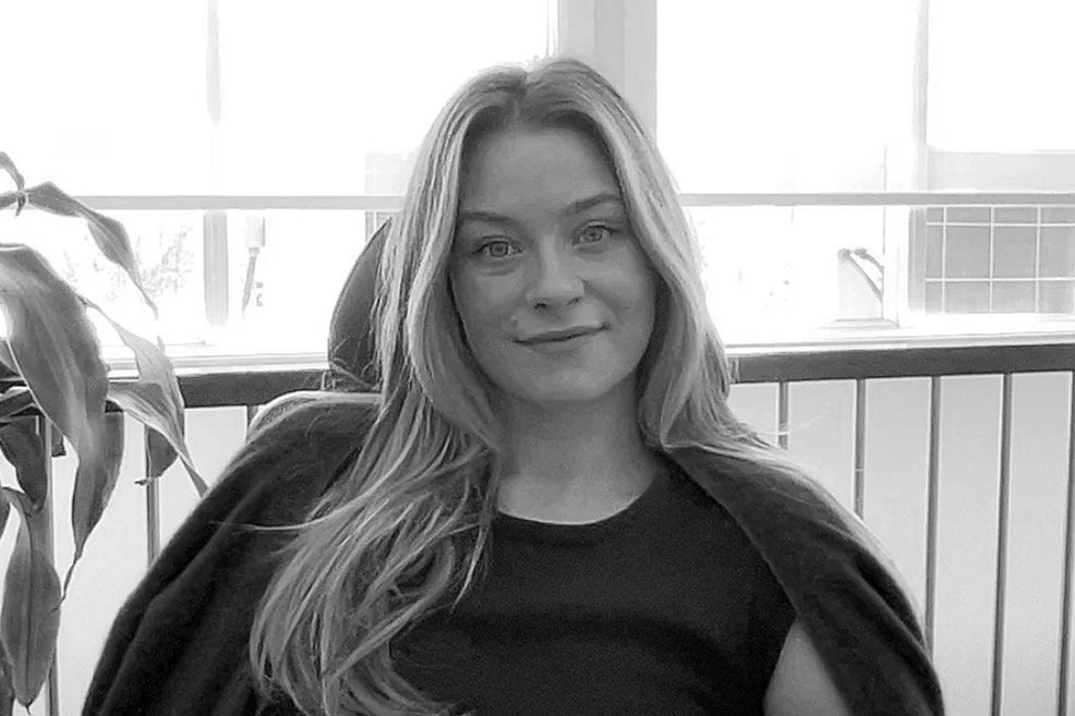 A young woman with long, light-colored hair, sitting indoors near a window, smiling slightly at the camera.