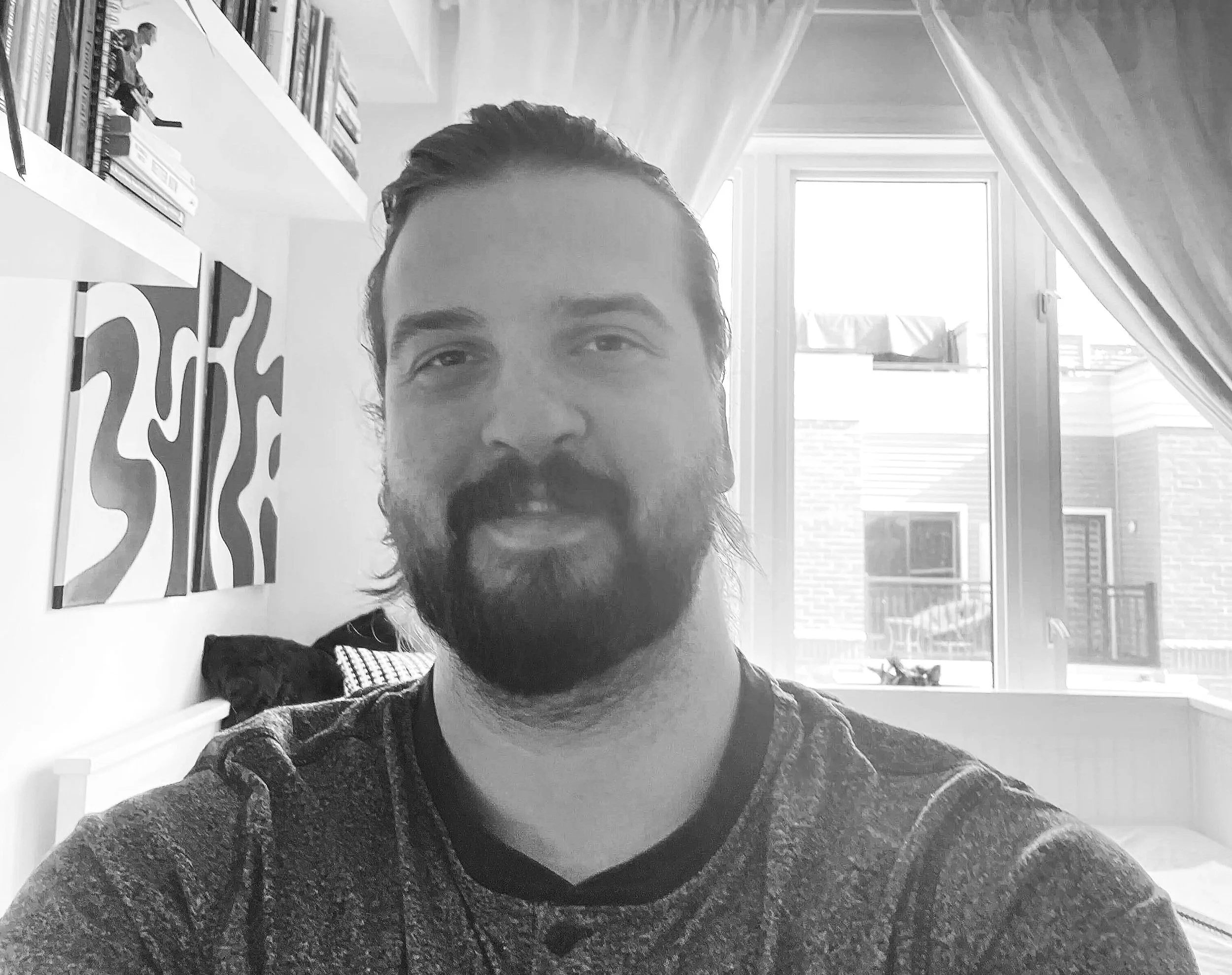 A man with a beard and mustache smiling in a room with a bookshelf and a window.