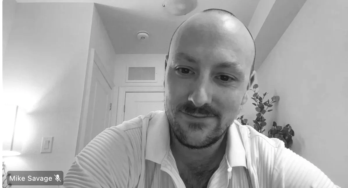 A man, smiling in a black and white photo, sitting in a room with a closed door, a plant, and a lamp visible in the background.