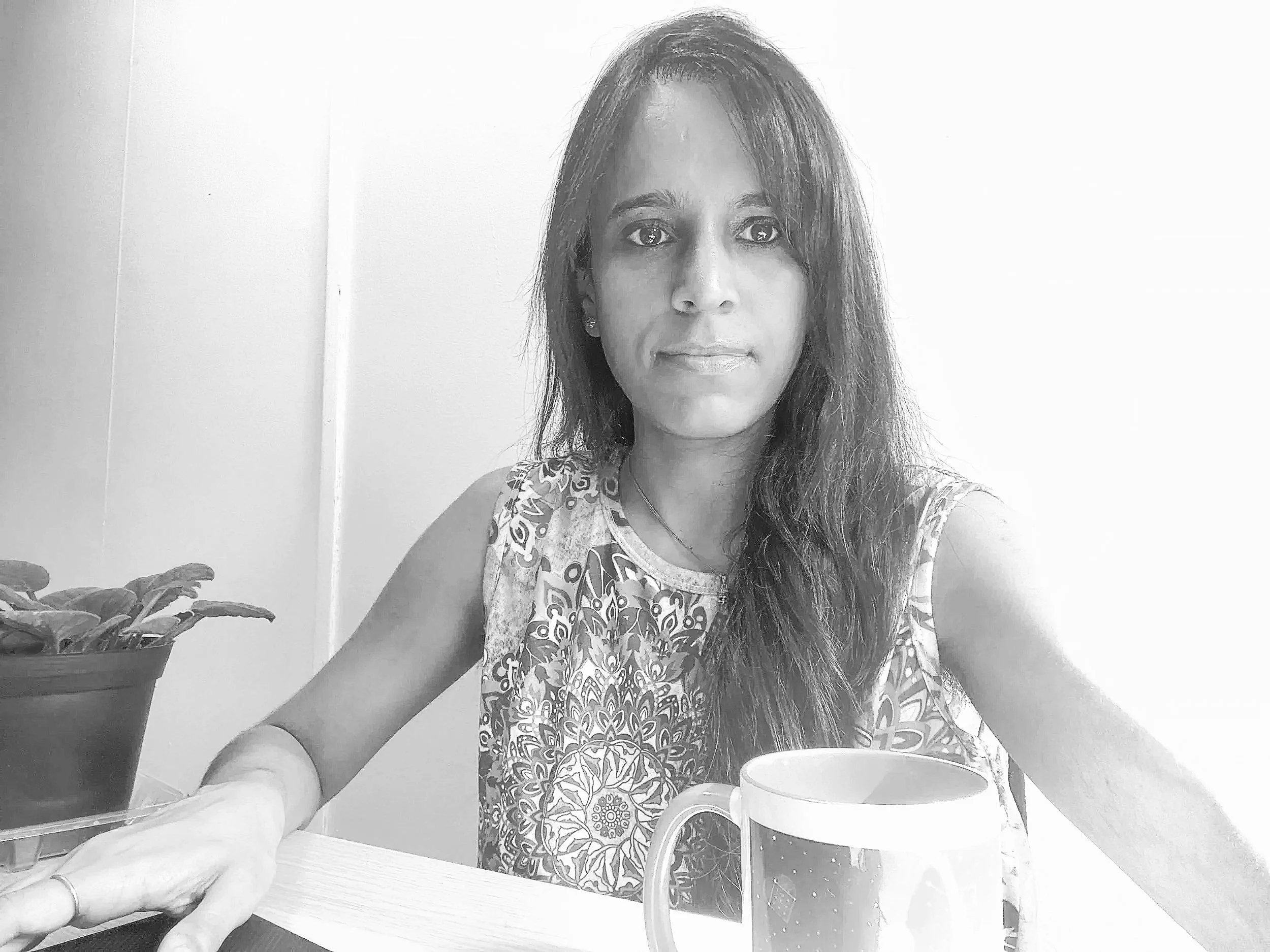 A woman with long hair sitting at a table with a mug in front of her, a potted plant to her side, in black and white.