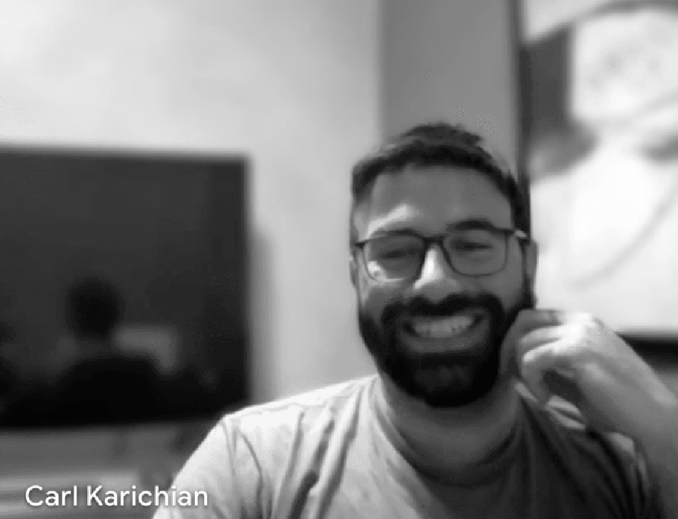 A smiling man with glasses and a beard sitting at a desk in front of a computer screen.