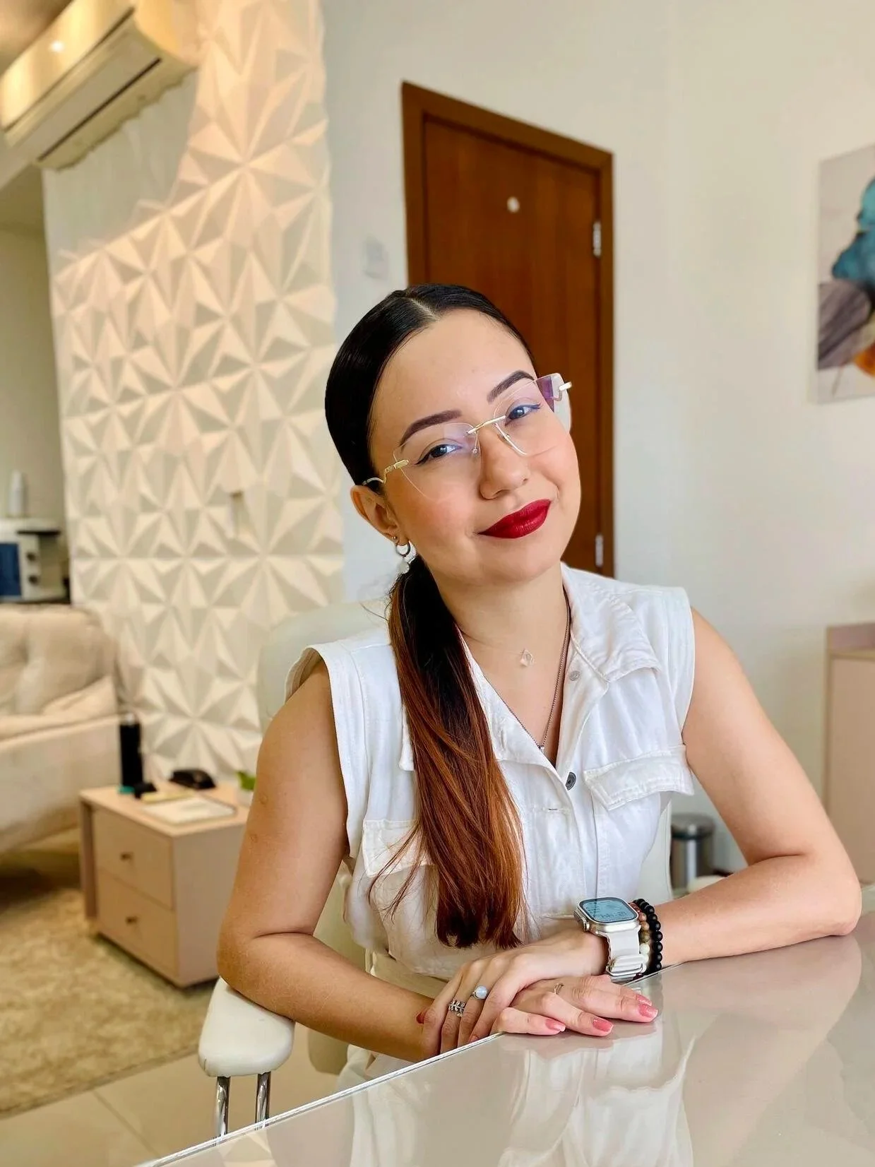 mulher com cabelo preto e castanho claro, óculos de grau, vestindo blusa branca, sentada em uma mesa de vidro com parede branca decorada ao fundo.