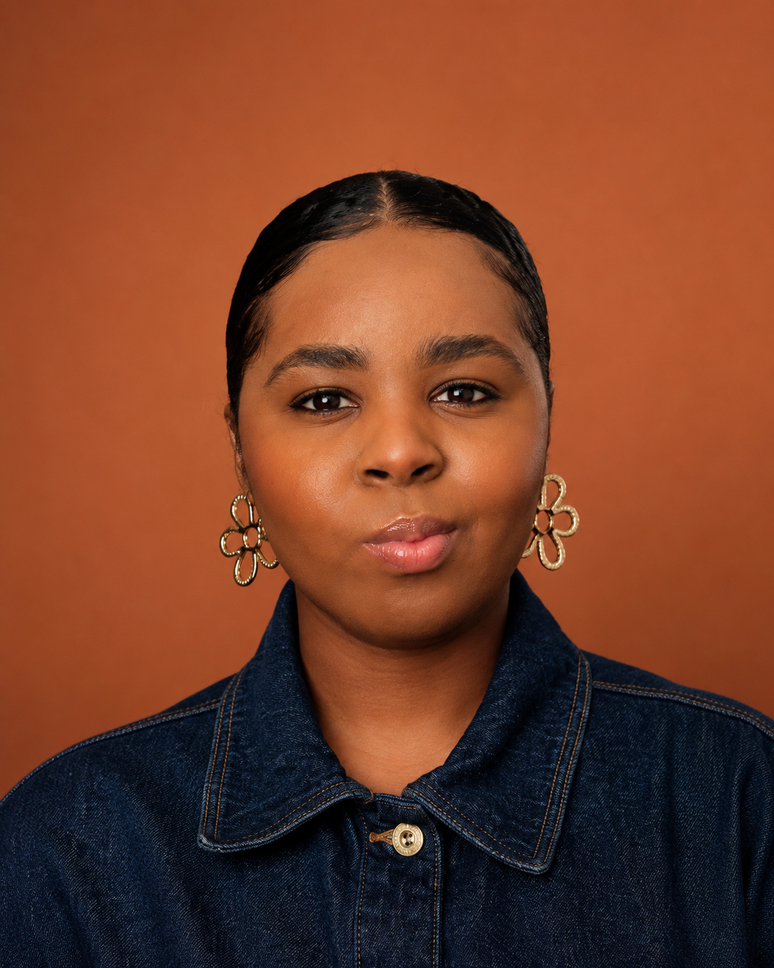 Portrait d'une jeune femme africaine avec des boucles d'oreilles en forme de fleurs, portant une veste en jean, contre un fond brun