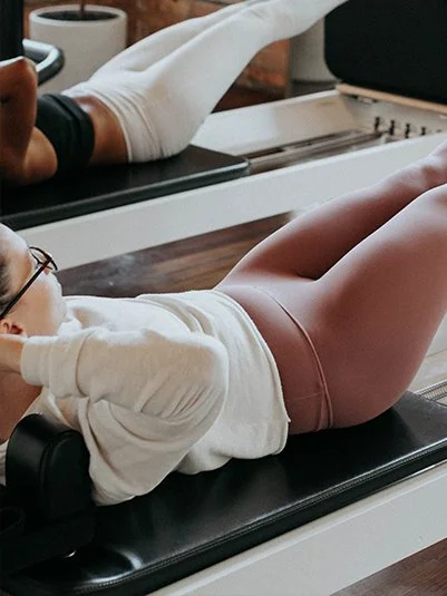 Child lying on pilates reformer machine in a fitness studio, wearing glasses, a white long-sleeve shirt, and pink shorts.