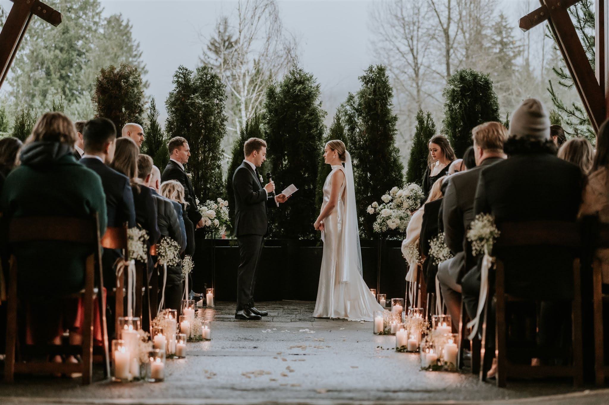 A wedding ceremony taking place outdoors with the bride and groom standing facing each other, surrounded by guests seated on both sides, with candles lining the aisle and trees in the background.