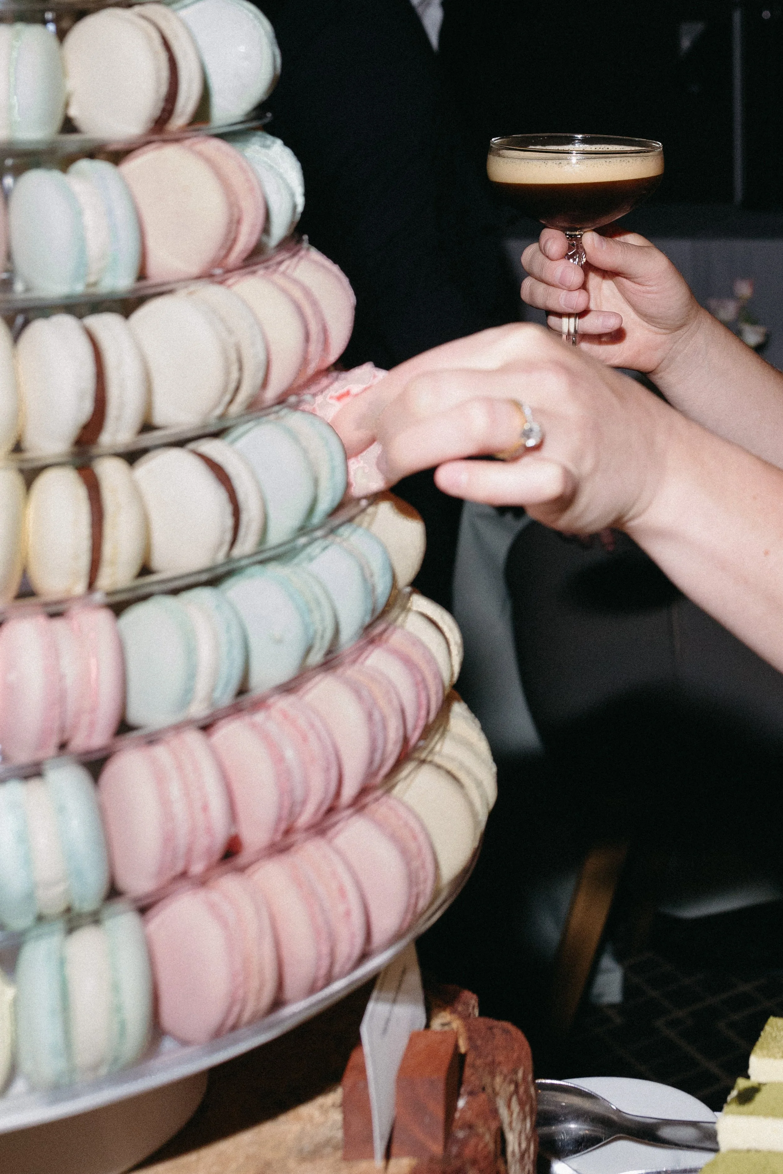 A person holding a champagne glass filled with a dark beverage, reaching for colorful macarons on a tiered display at a dessert table.