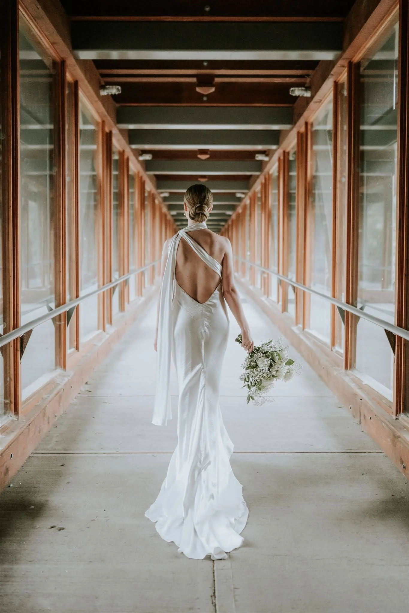 A bride in a backless white wedding dress holding a bouquet walking down a glass hallway with wooden frames.