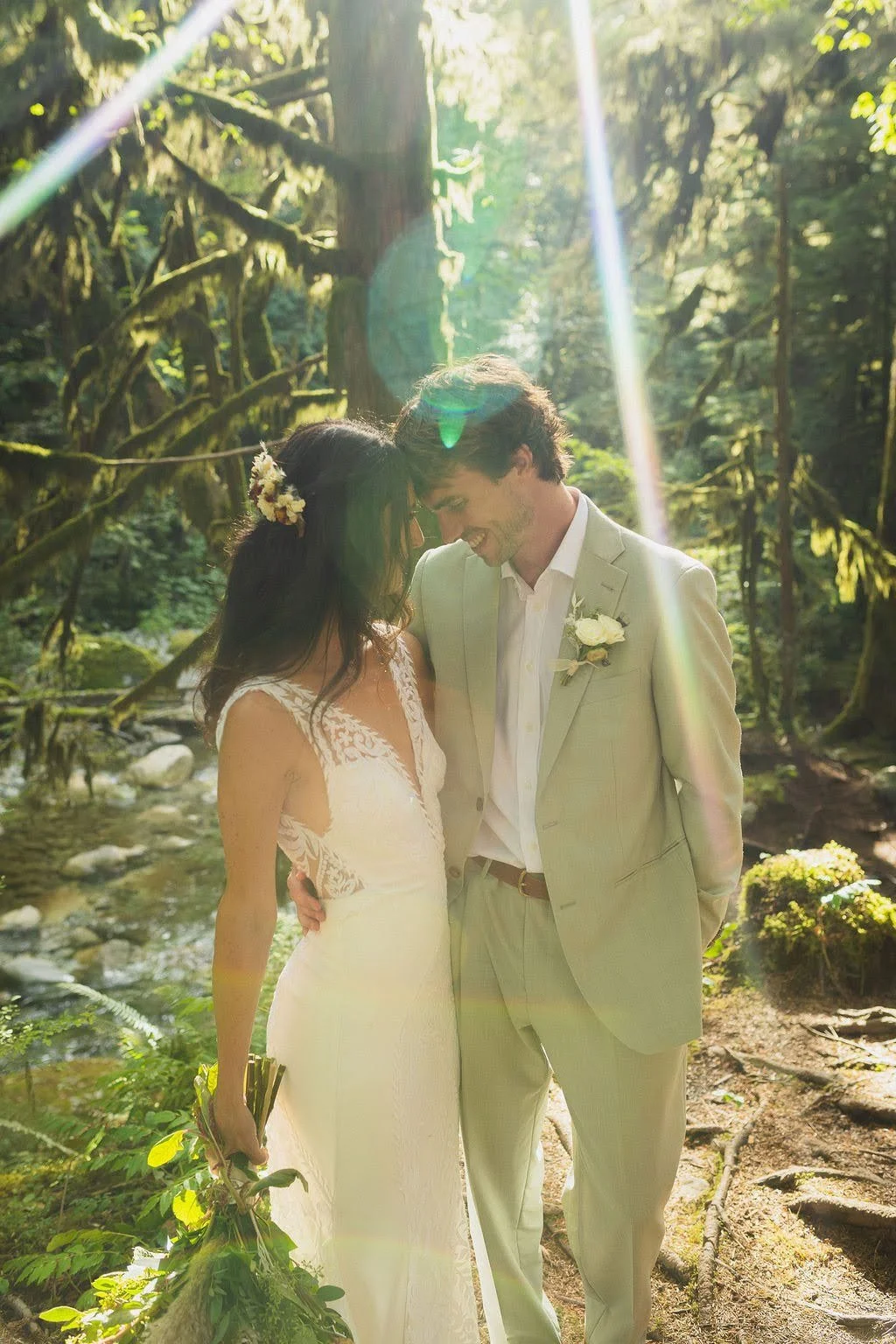 A bride and groom standing close together in a forest with sunlight streaming through trees, smiling and touching foreheads. The bride holds a bouquet, and the groom has a boutonnière on his suit.