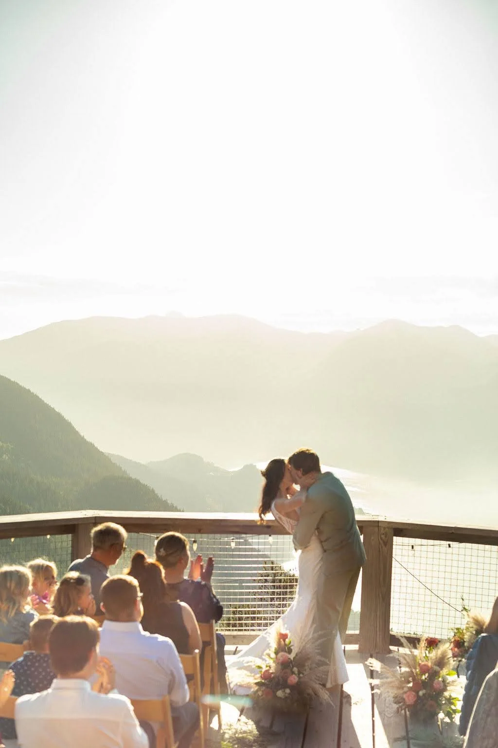 A newlywed couple sharing a kiss during their outdoor wedding ceremony, surrounded by guests, with mountains and a scenic landscape in the background at sunset.