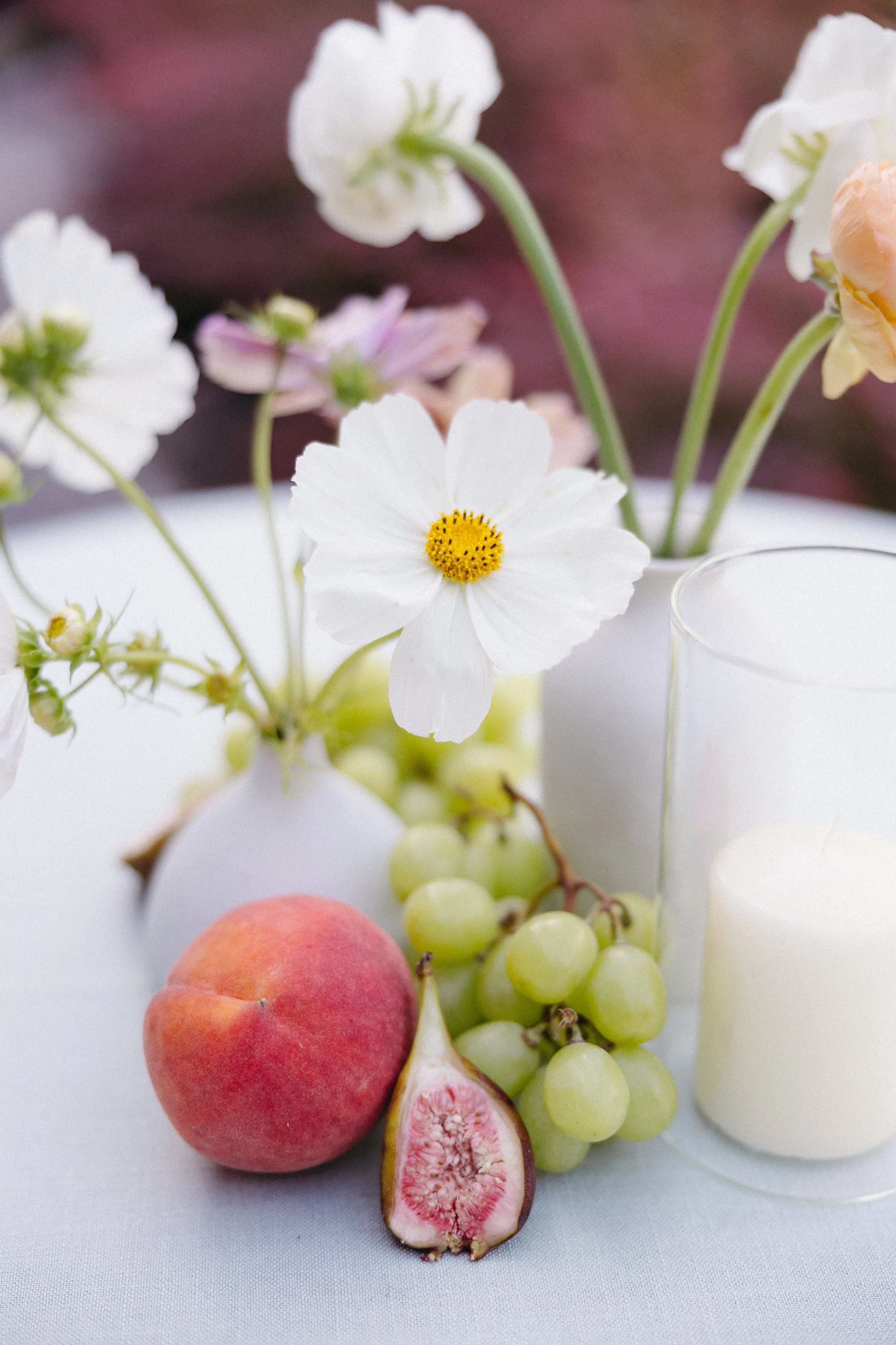 A still life of white and pink flowers in vases, a peach, a sliced fig, green grapes, and candles on a white surface.