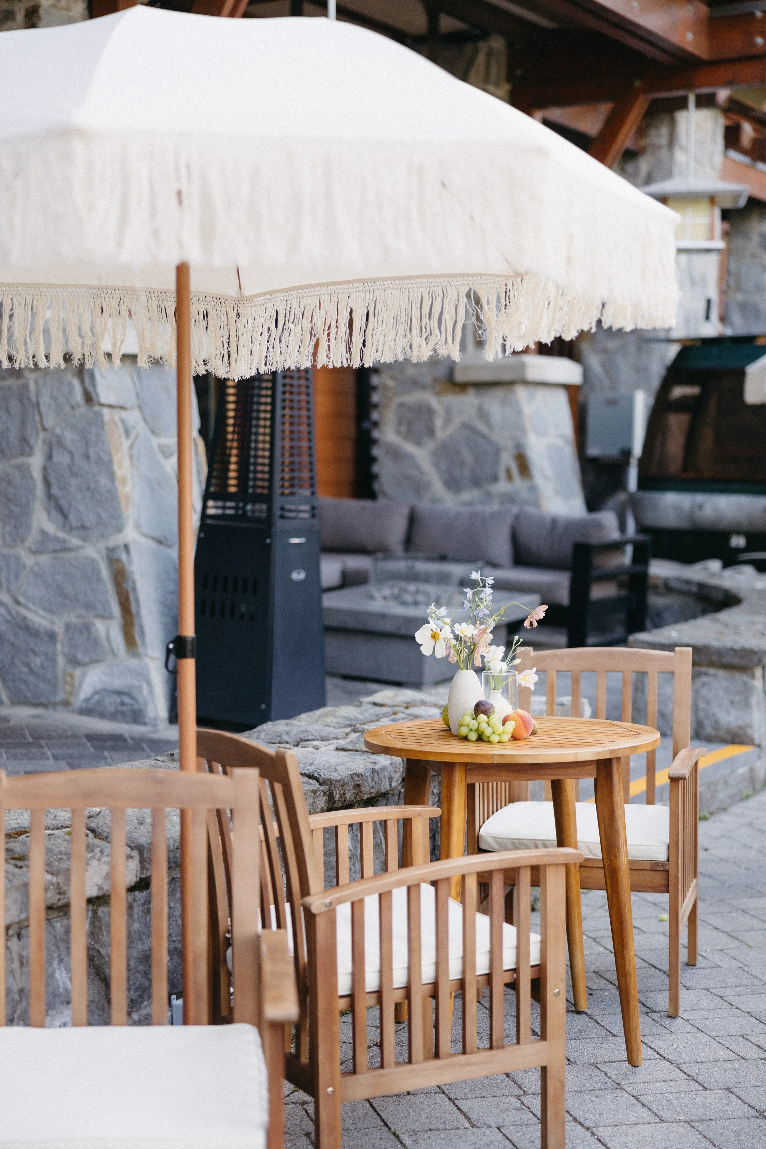 Outdoor patio with wooden chairs, a round wooden table with a vase of flowers and fruit, and a large cream sun umbrella. Stone wall and patio furniture in the background.