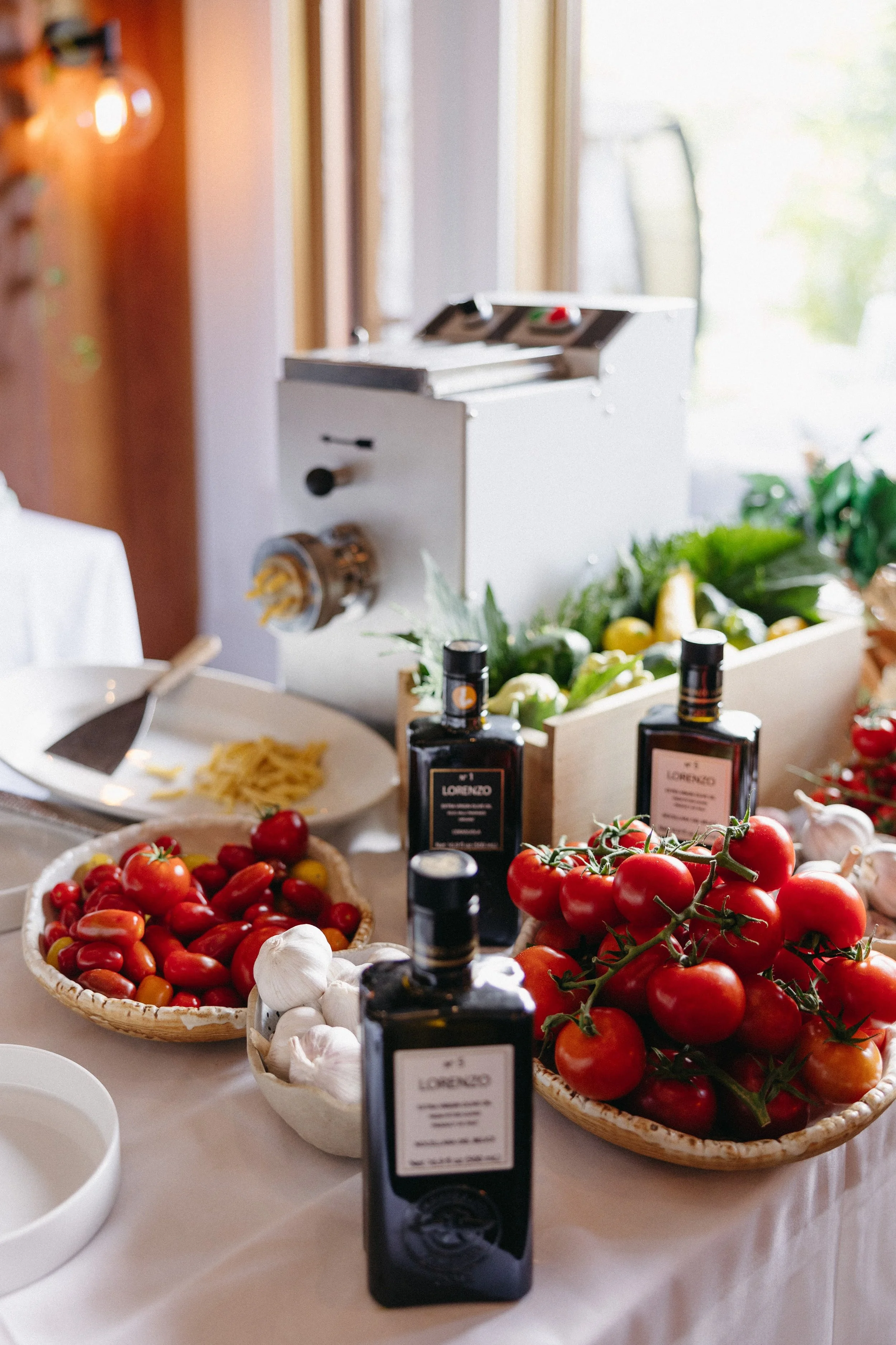 Fresh tomatoes, garlic, and bottles of olive oil on a white table near a pasta machine with vegetables and greens in the background.