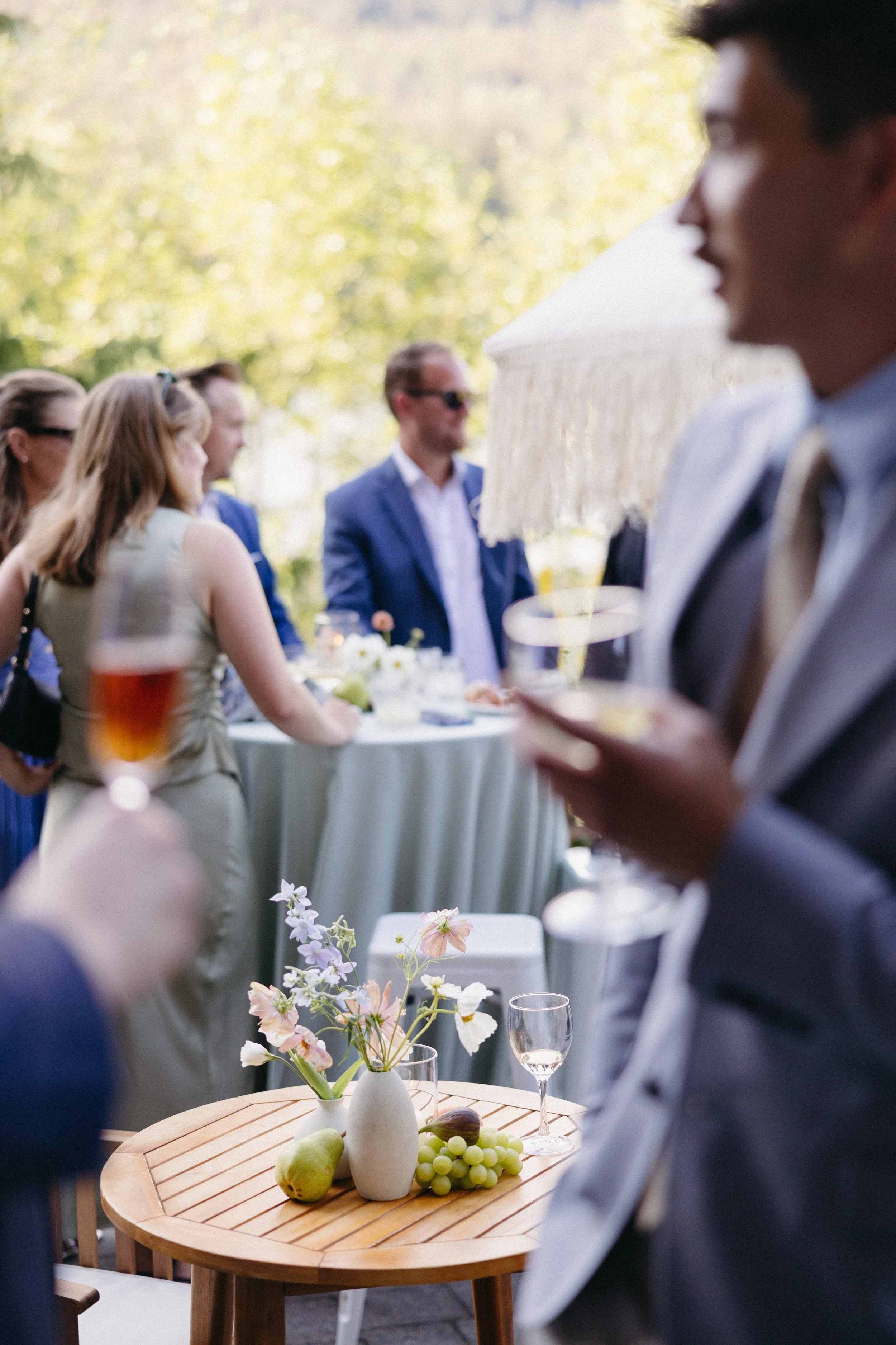 People gathered at an outdoor event with tables decorated with flowers, fruit, and drinks, under a white umbrella with trees and greenery in the background.