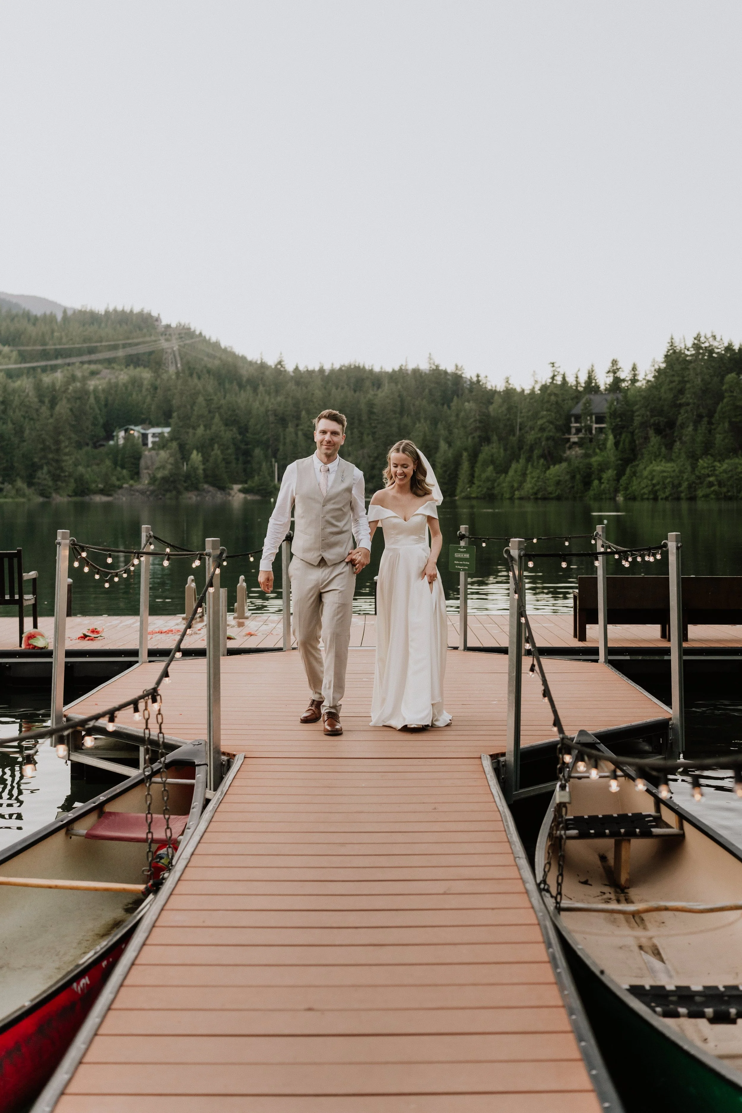 A bride and groom walking hand in hand on a wooden dock by a lake, surrounded by trees and mountains.