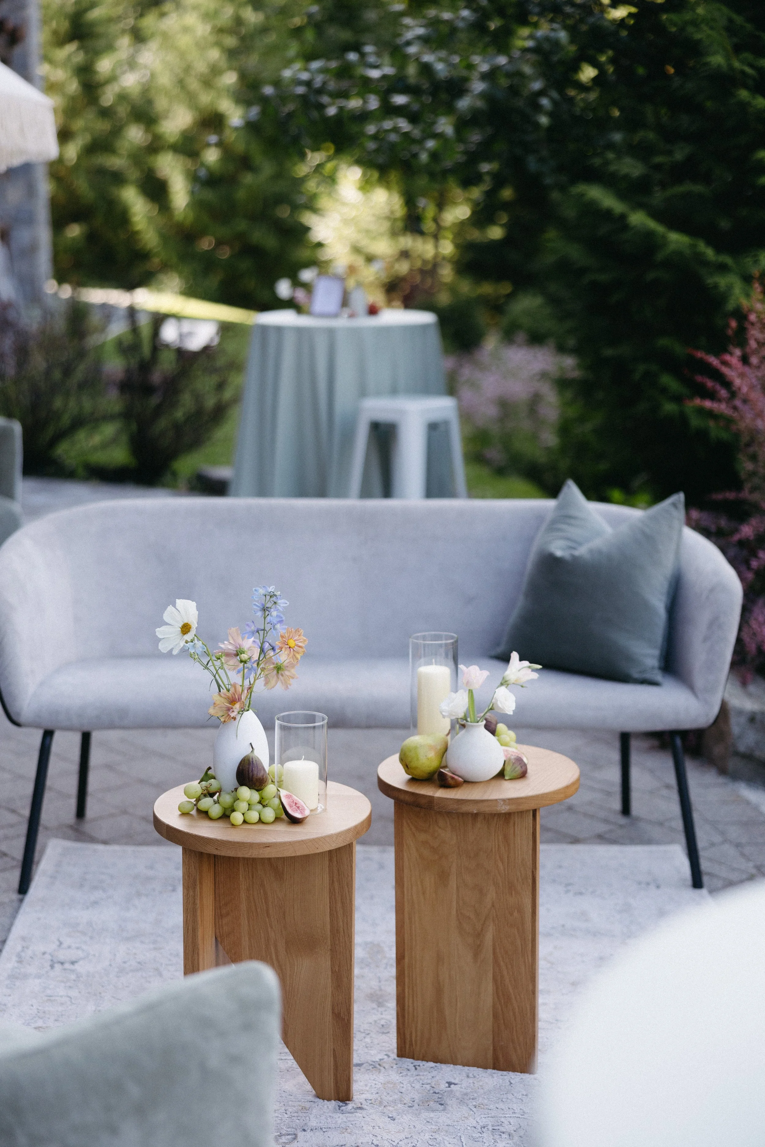 Outdoor seating area with a beige couch, a gray pillow, and two wooden tables decorated with vases of flowers, candles, grapes, and pears, set on a patio surrounded by greenery.