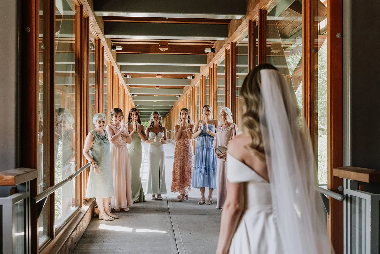 A bride in a white wedding dress with a veil faces a group of women in pastel-colored dresses in a wooden corridor with large windows.