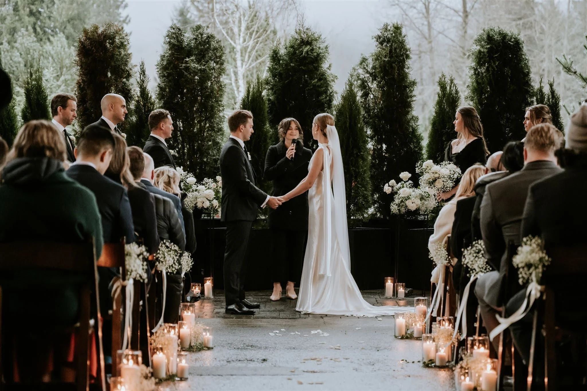 Outdoor wedding ceremony with a bride and groom holding hands, exchanging vows, under a row of tall green trees, surrounded by guests seated on both sides with candles and flowers.