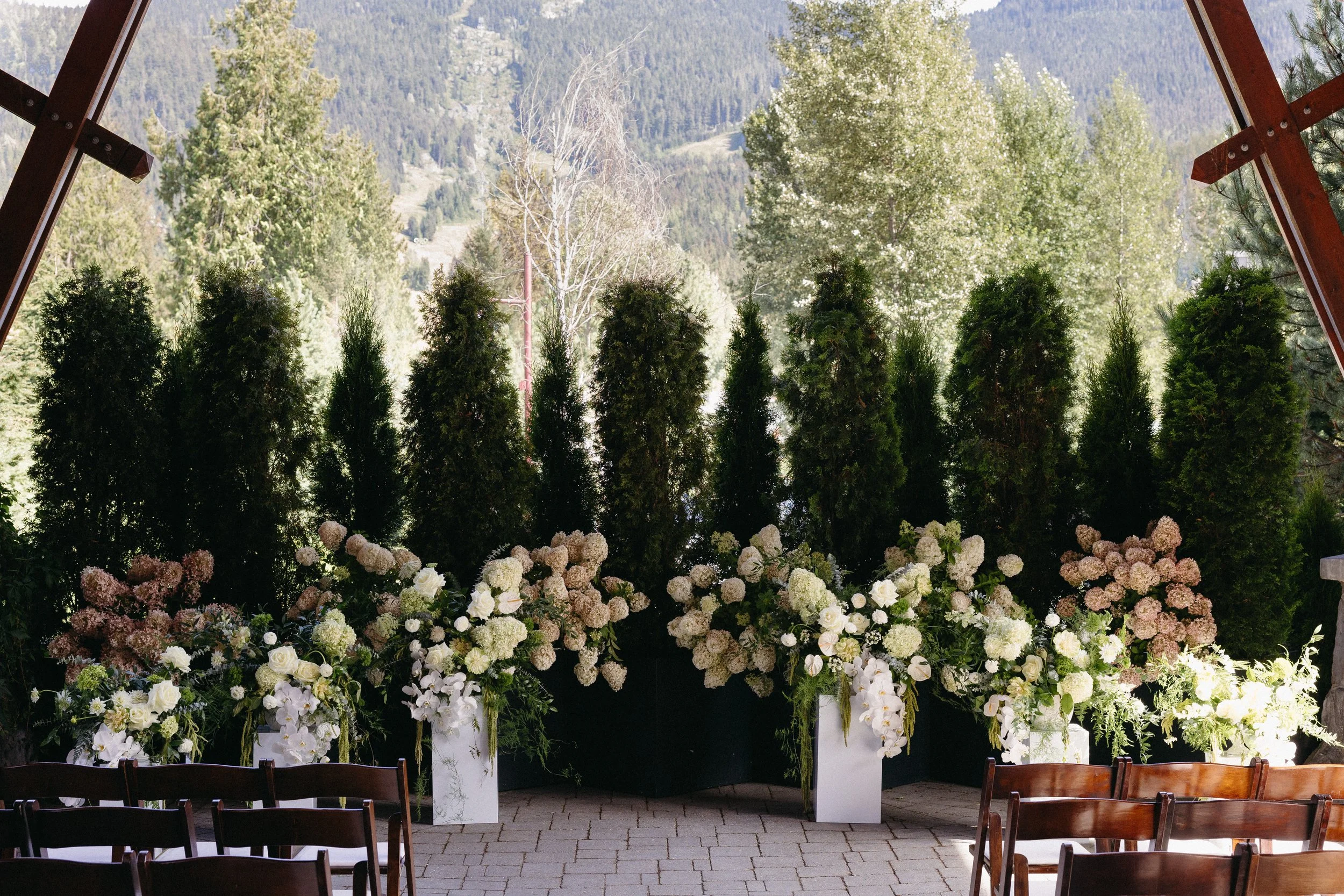 Indoor wedding altar with floral arrangements and white chairs, outdoor mountain view in background.