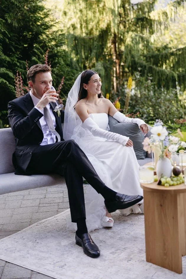 A bride in a white wedding dress and veil sitting on a gray outdoor sofa, smiling, with a groom in a black suit and white shirt sitting beside her, drinking a glass of wine, in a lush garden setting with greenery and flowers.