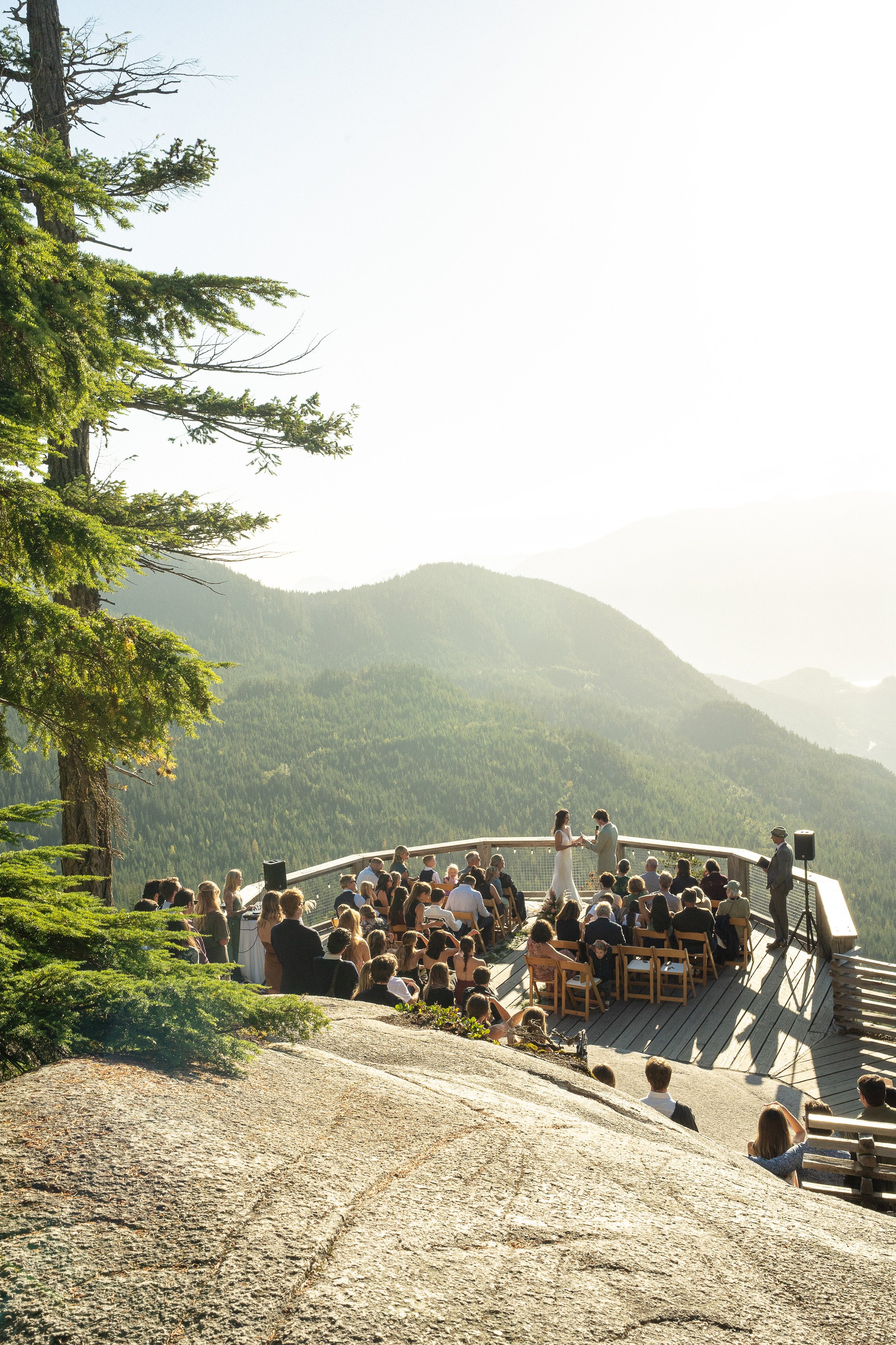 A wedding ceremony taking place outdoors on a wooden deck overlooking a mountain landscape with trees.